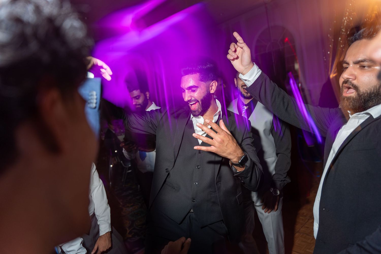 Wedding guests dancing and celebrating with excitement on a colorfully lit dance floor at a reception.