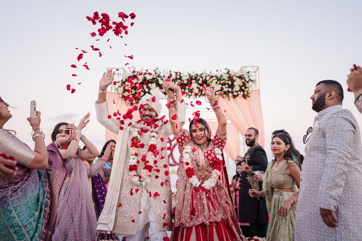 Red rose petals shower down during a festive celebration with people gathered around a floral backdrop.