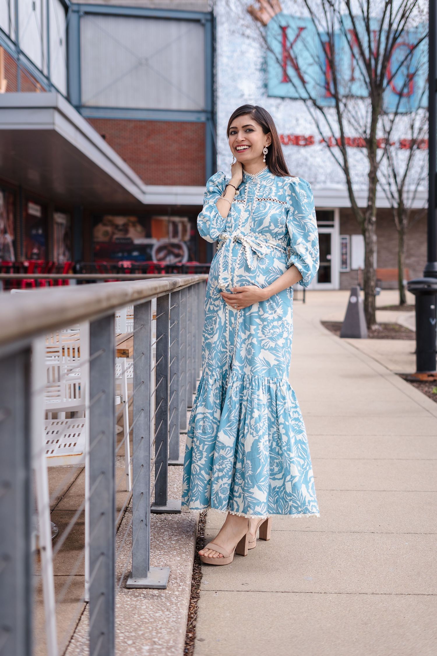 Expectant mother wearing a flowing blue and white paisley maxi dress stands on an outdoor sidewalk next to a railing.