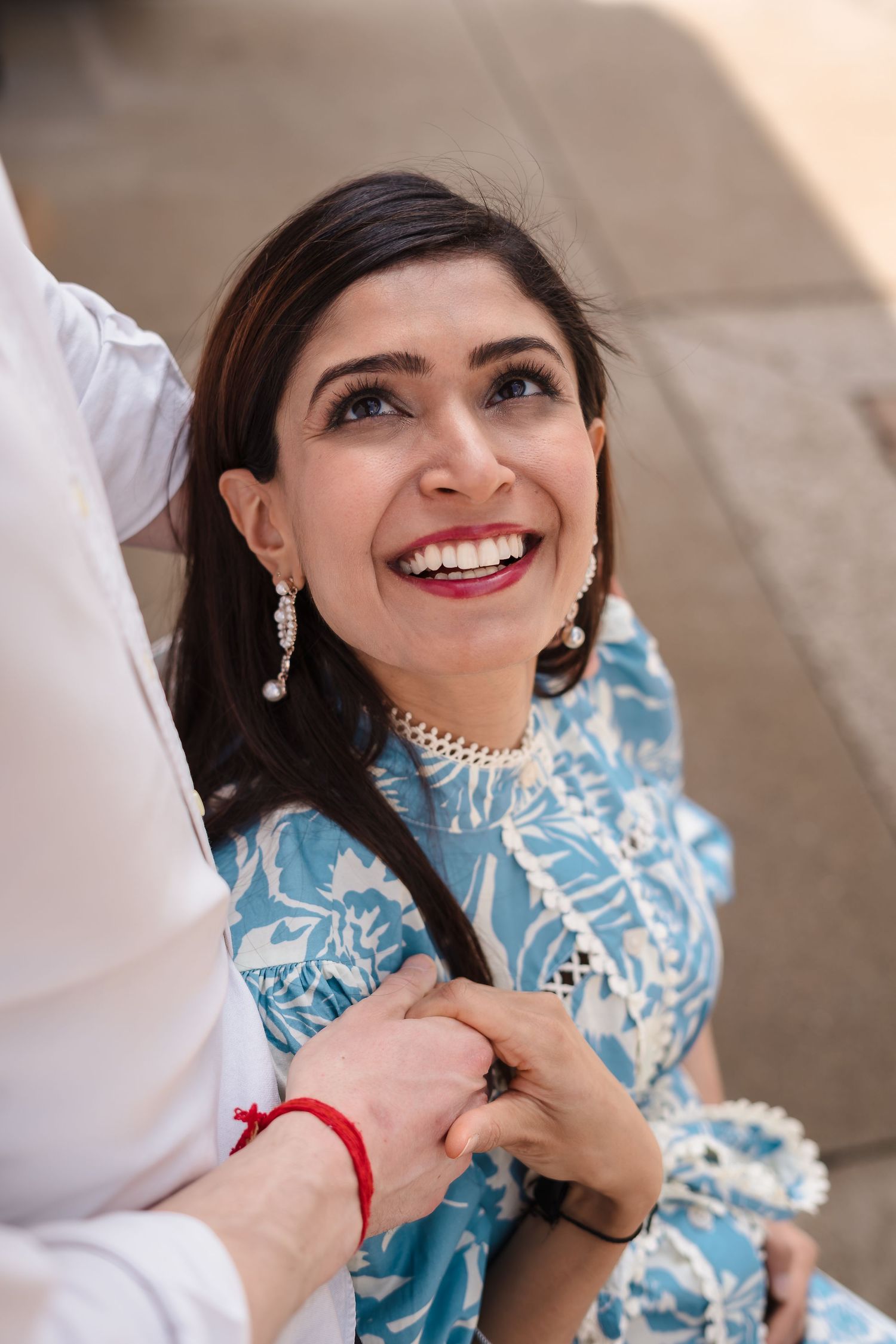 A person wearing a blue and white patterned dress and dangling earrings smiles while holding hands with someone.