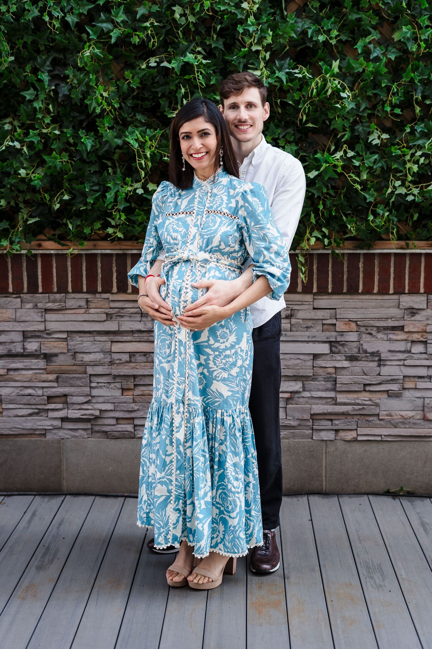 A couple stands together against a stone wall and greenery backdrop, with a blue and white patterned maternity dress.