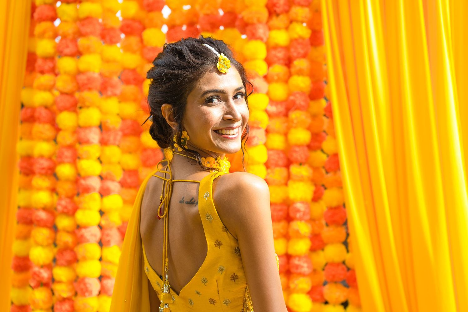 Woman in yellow backless formal dress poses against bright orange and yellow backdrop with floral decor.