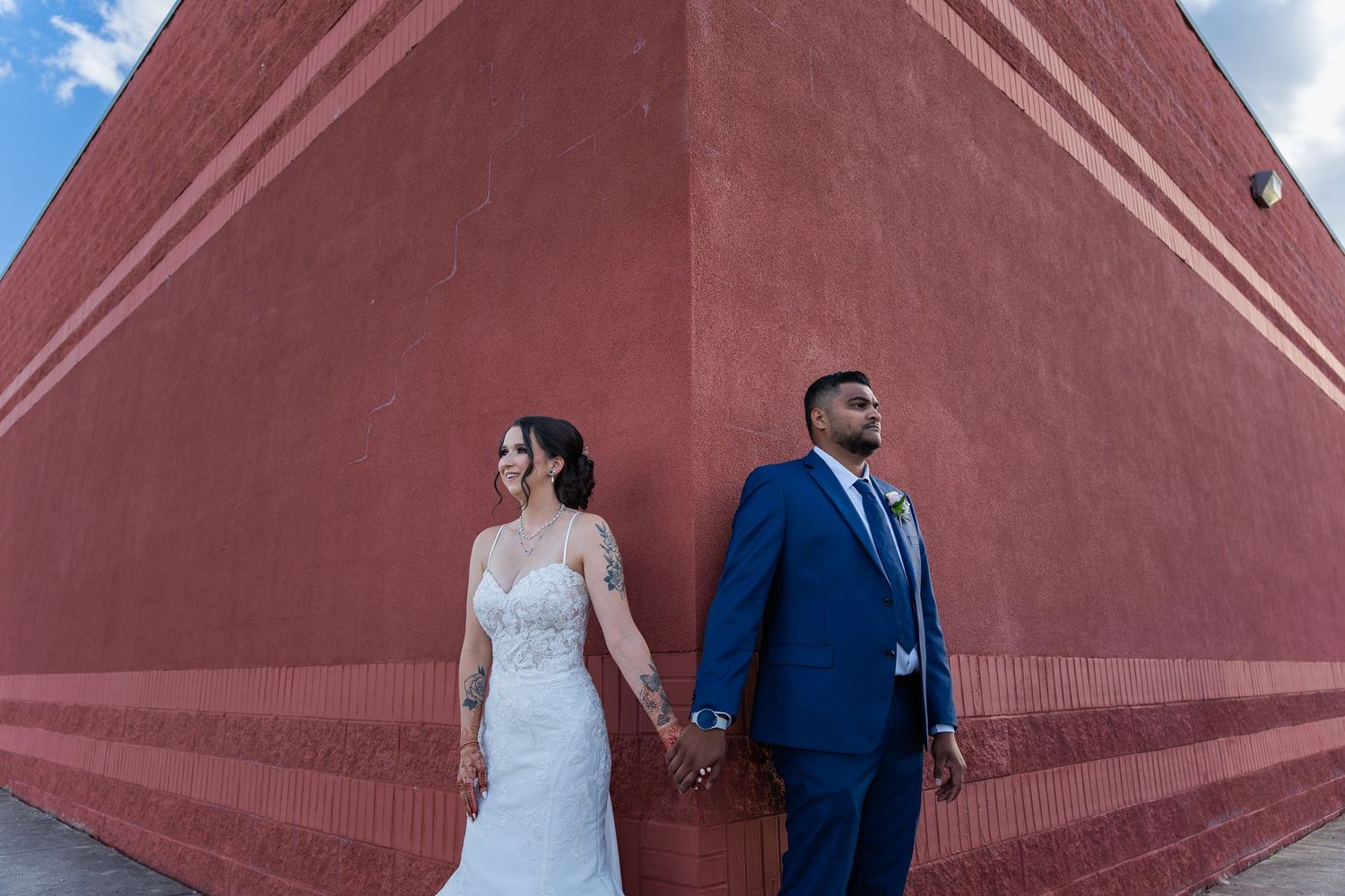 Bride in white gown and groom in blue suit hold hands against angular terracotta wall.