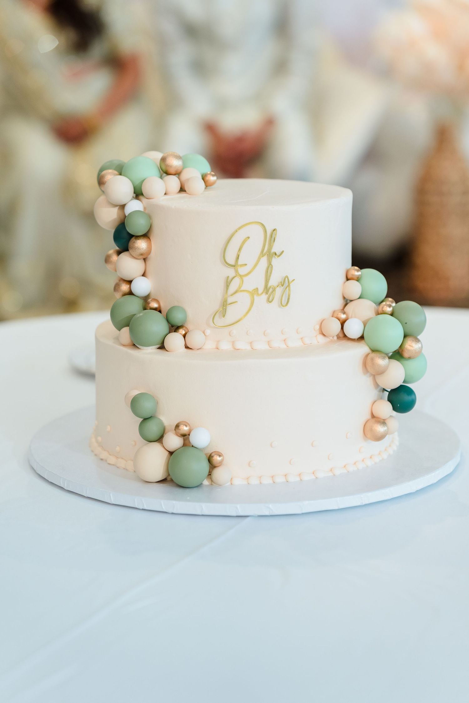 Elegant two-tier white cake decorated with mint green and gold balloon garland and 'Oh Baby' text.