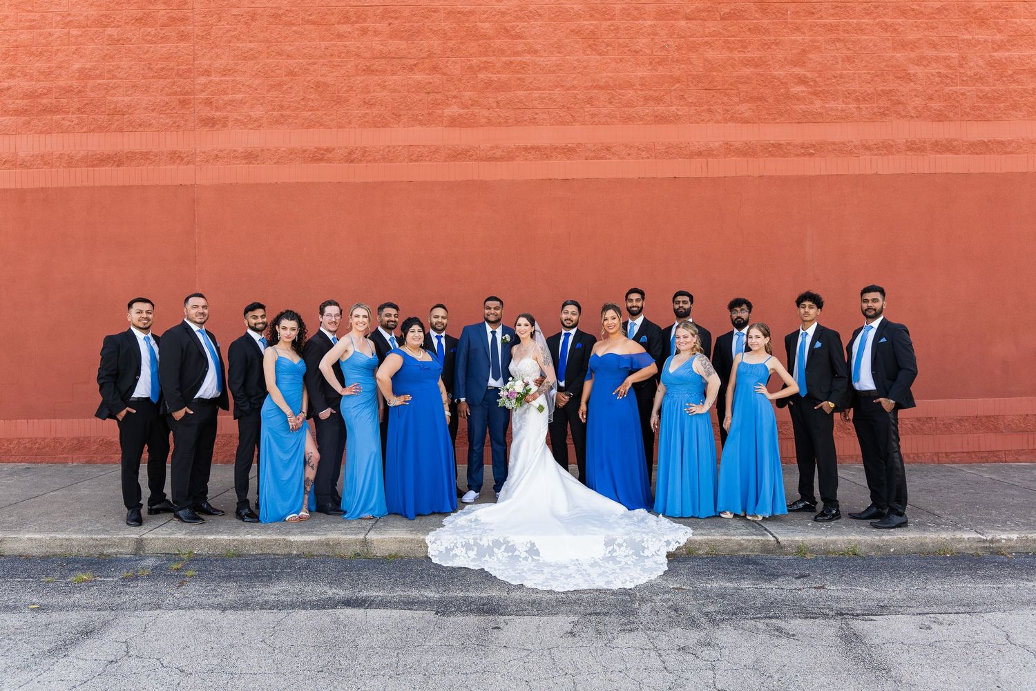 Wedding party in blue and black attire poses formally against red wall.