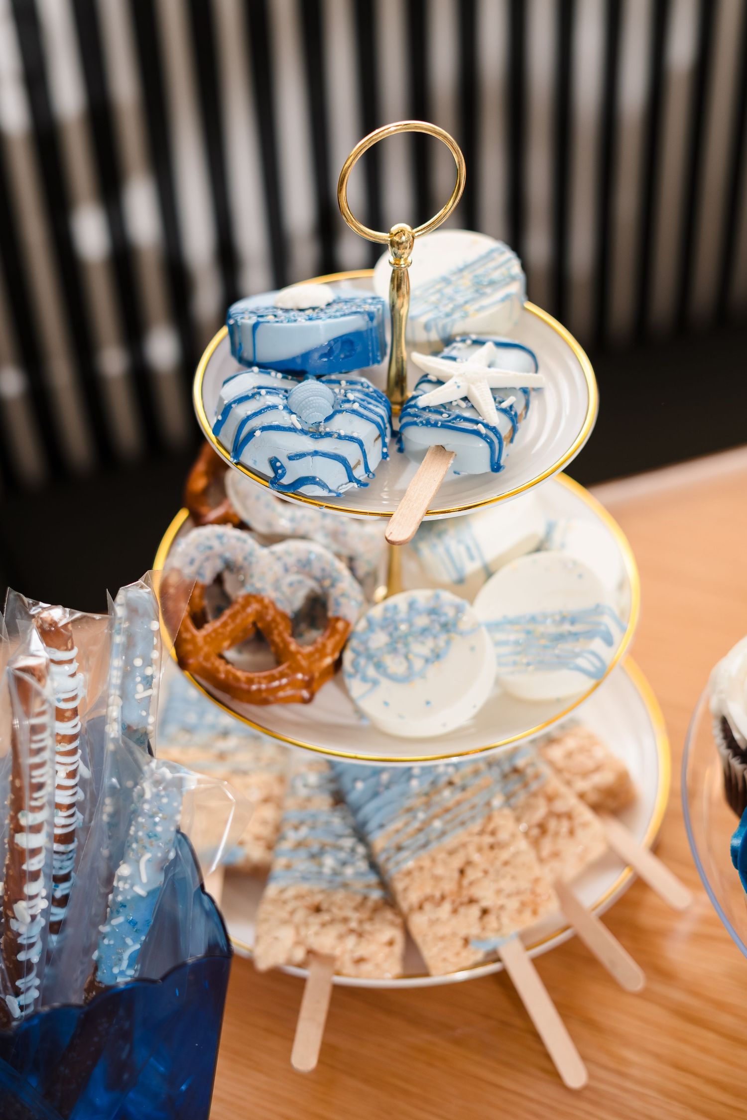 A tiered serving stand displays blue and white decorated cookies and treats with a glass of blue drink on the side.
