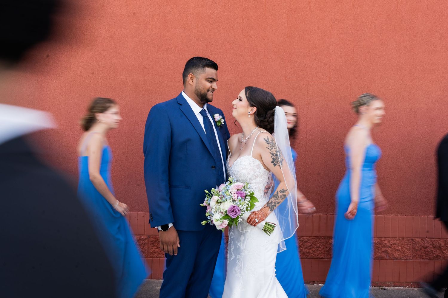 Newlyweds pose with bridesmaids in blue dresses against a coral colored wall.