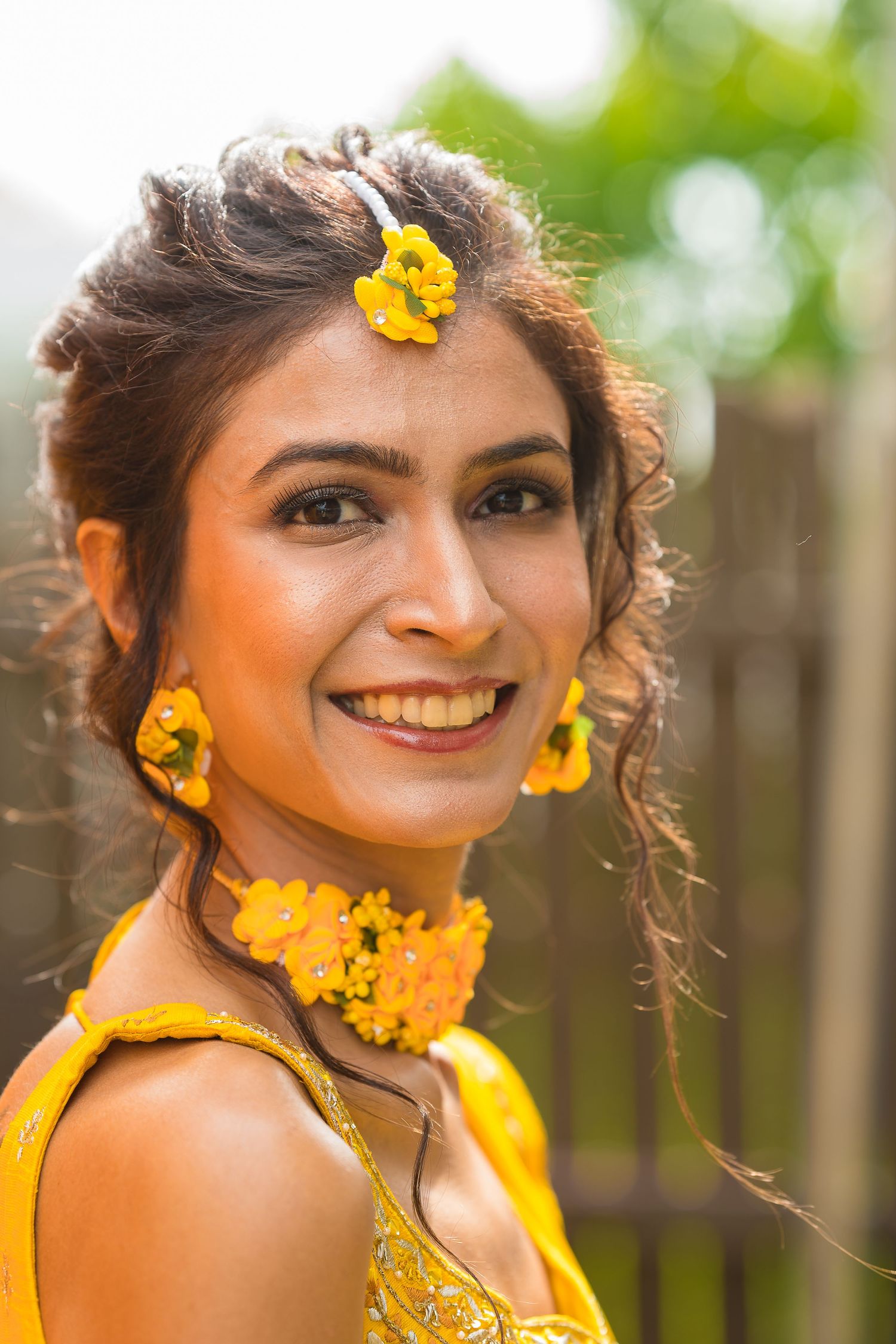 A woman wearing yellow floral accessories smiles brightly in natural outdoor lighting.
