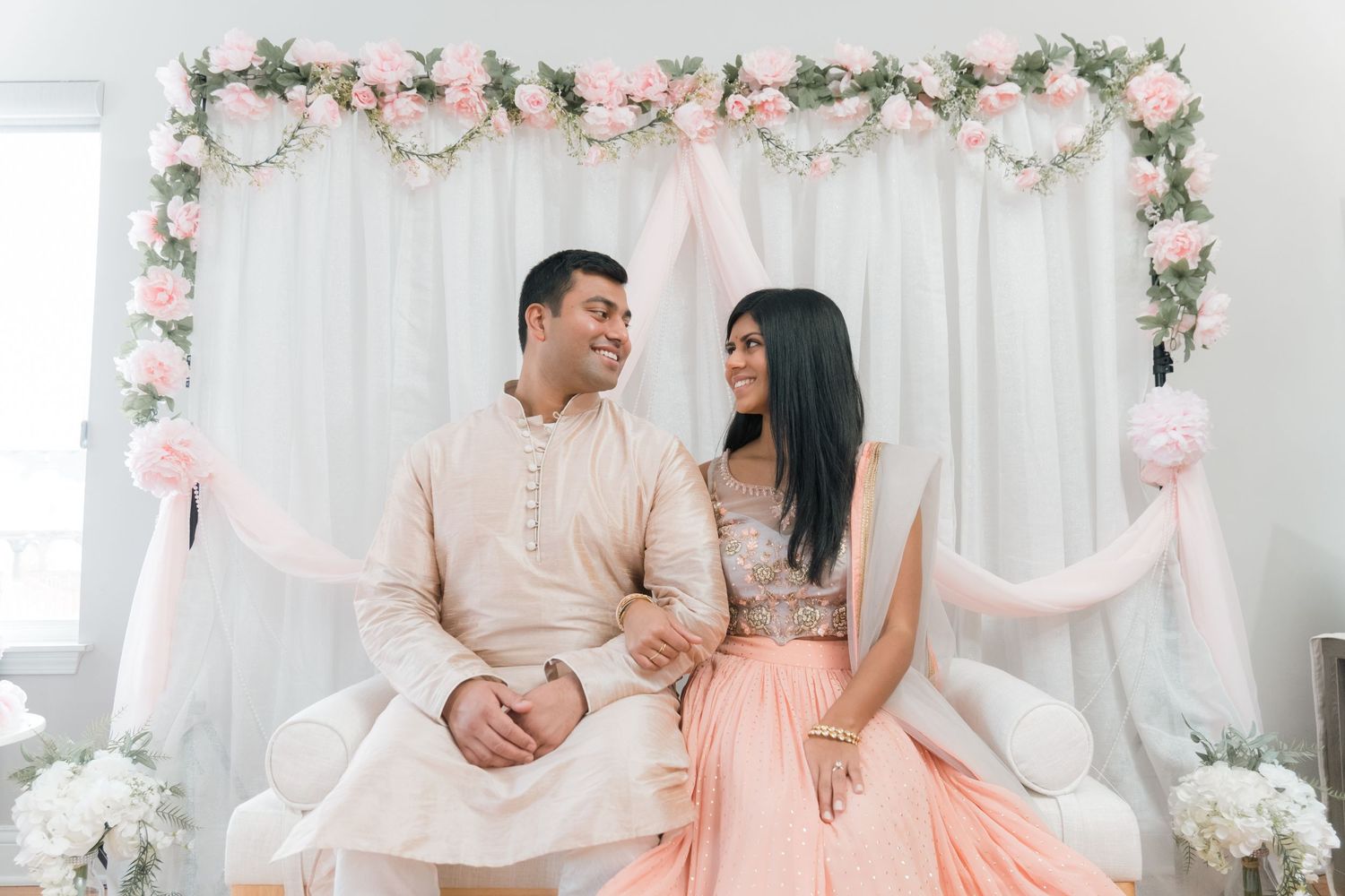 A couple in pastel traditional attire poses under a floral arch with pink and white decorations.