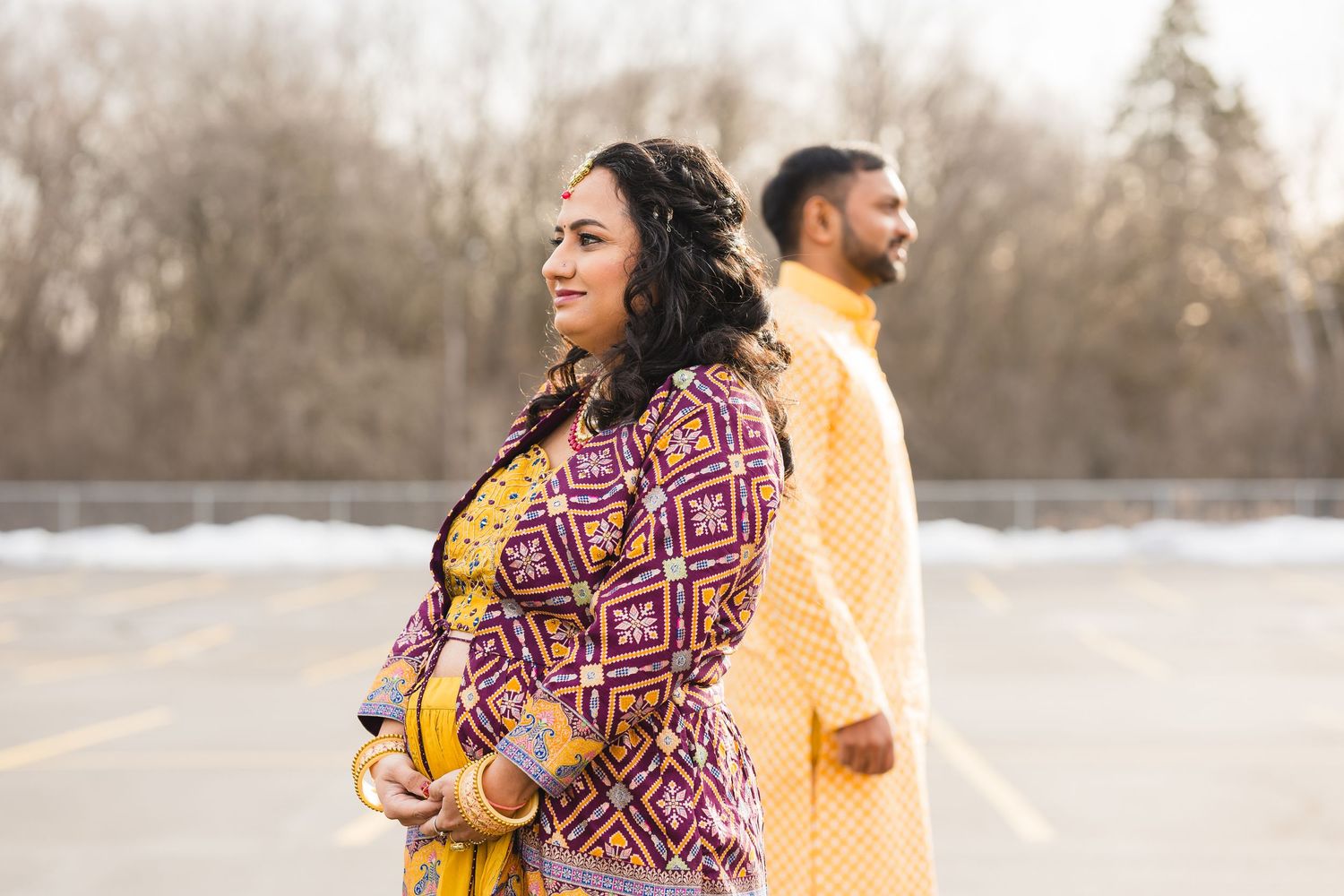 A couple in colorful traditional outfits poses back-to-back outdoors during winter.