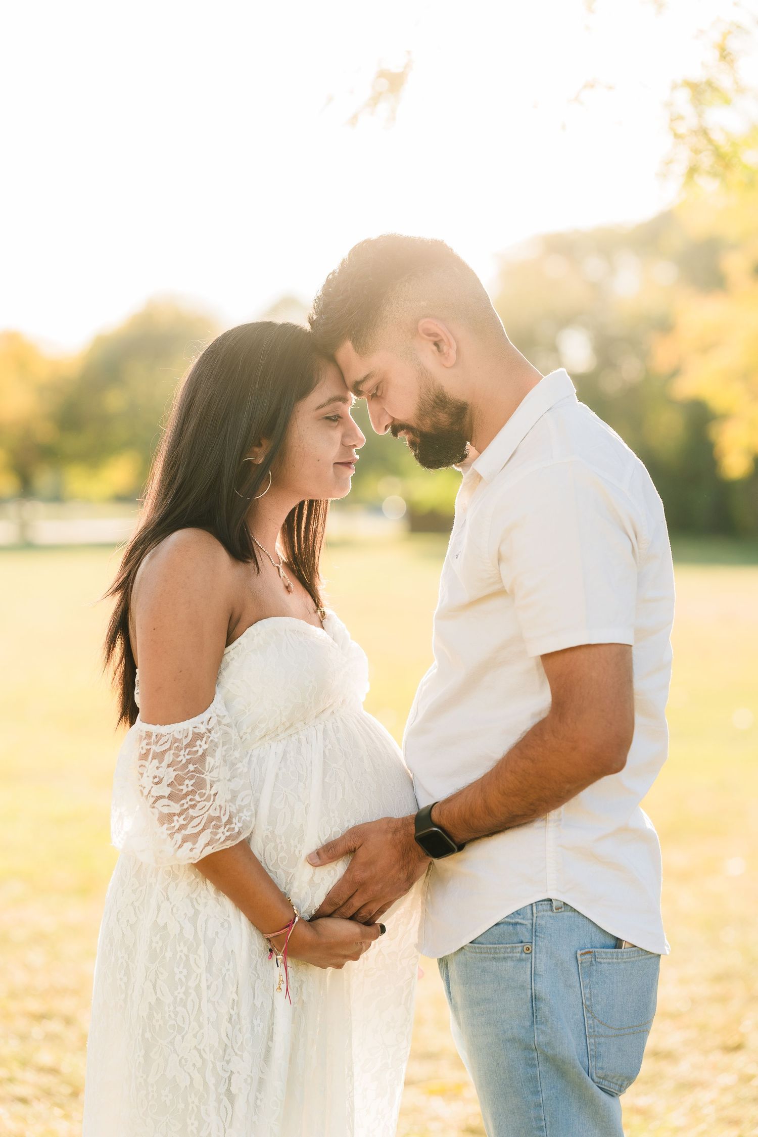 A couple shares an intimate moment at sunset in a park while wearing white and embracing a pregnant belly.