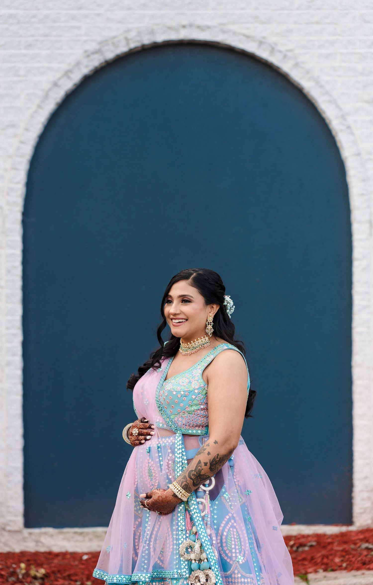 Person posing in blue and pink traditional Indian outfit against arched blue wall backdrop.
