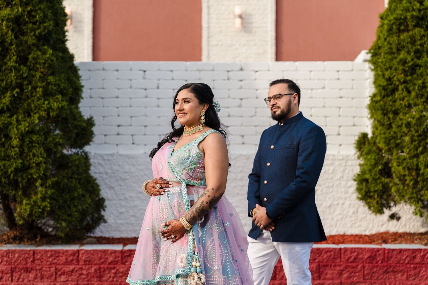 A couple stands in front of a white brick wall with greenery wearing elegant Indo-western outfits.