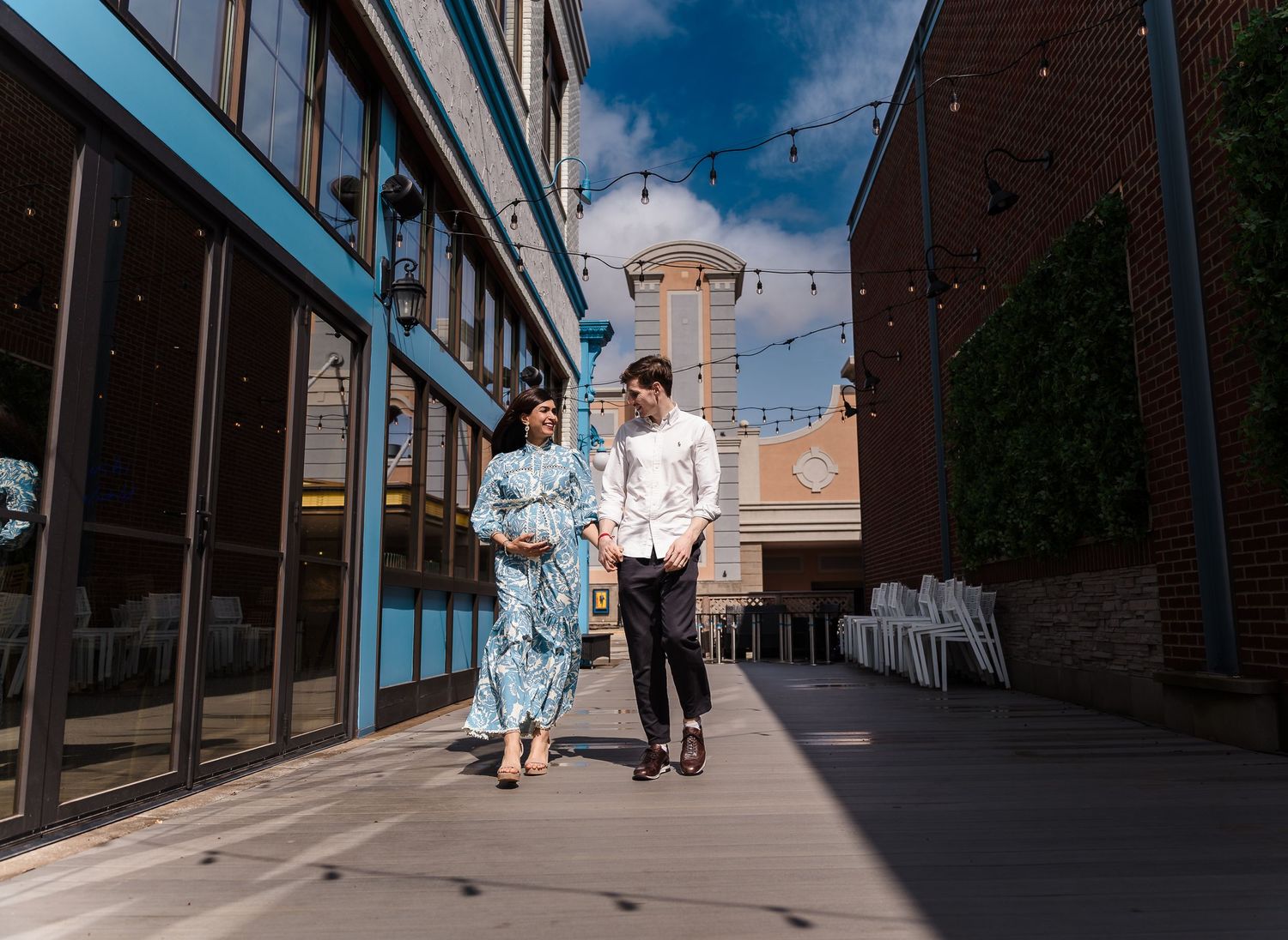 A couple walking together on an urban sidewalk with string lights and buildings in the background on a sunny day.