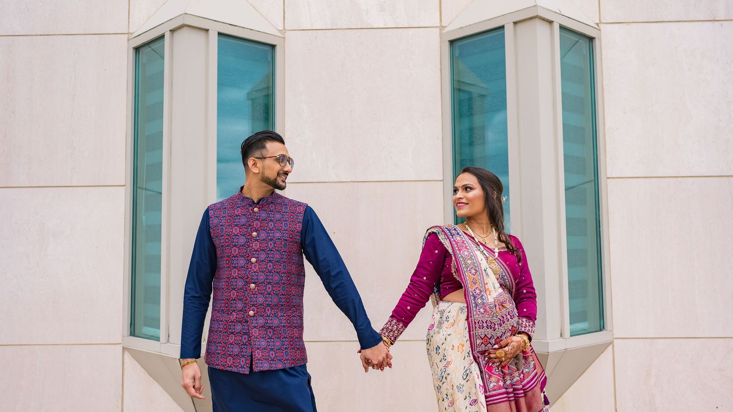 Couple in traditional Indian attire holding hands against modern architectural backdrop.