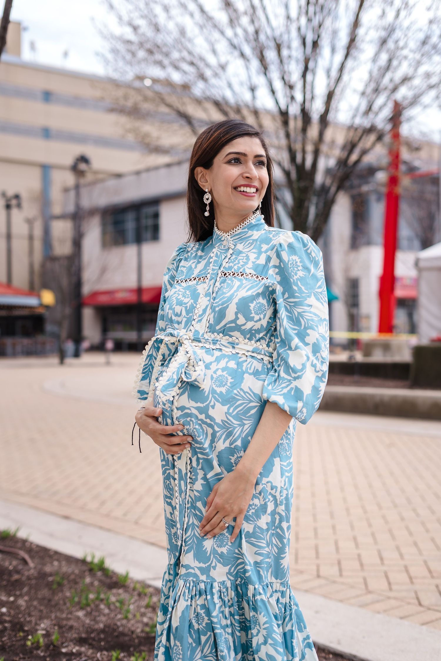 Elegant light blue and white patterned maternity dress with balloon sleeves and ruffled hem shown on a brick sidewalk.
