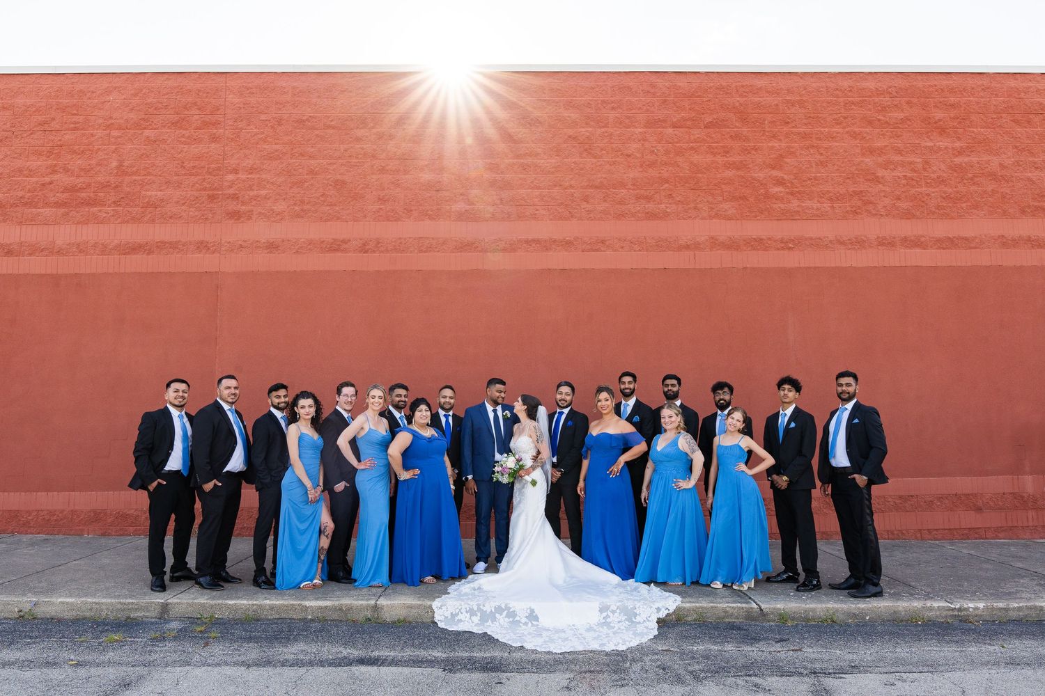 Wedding party in blue and white formal attire posed against red wall backdrop.