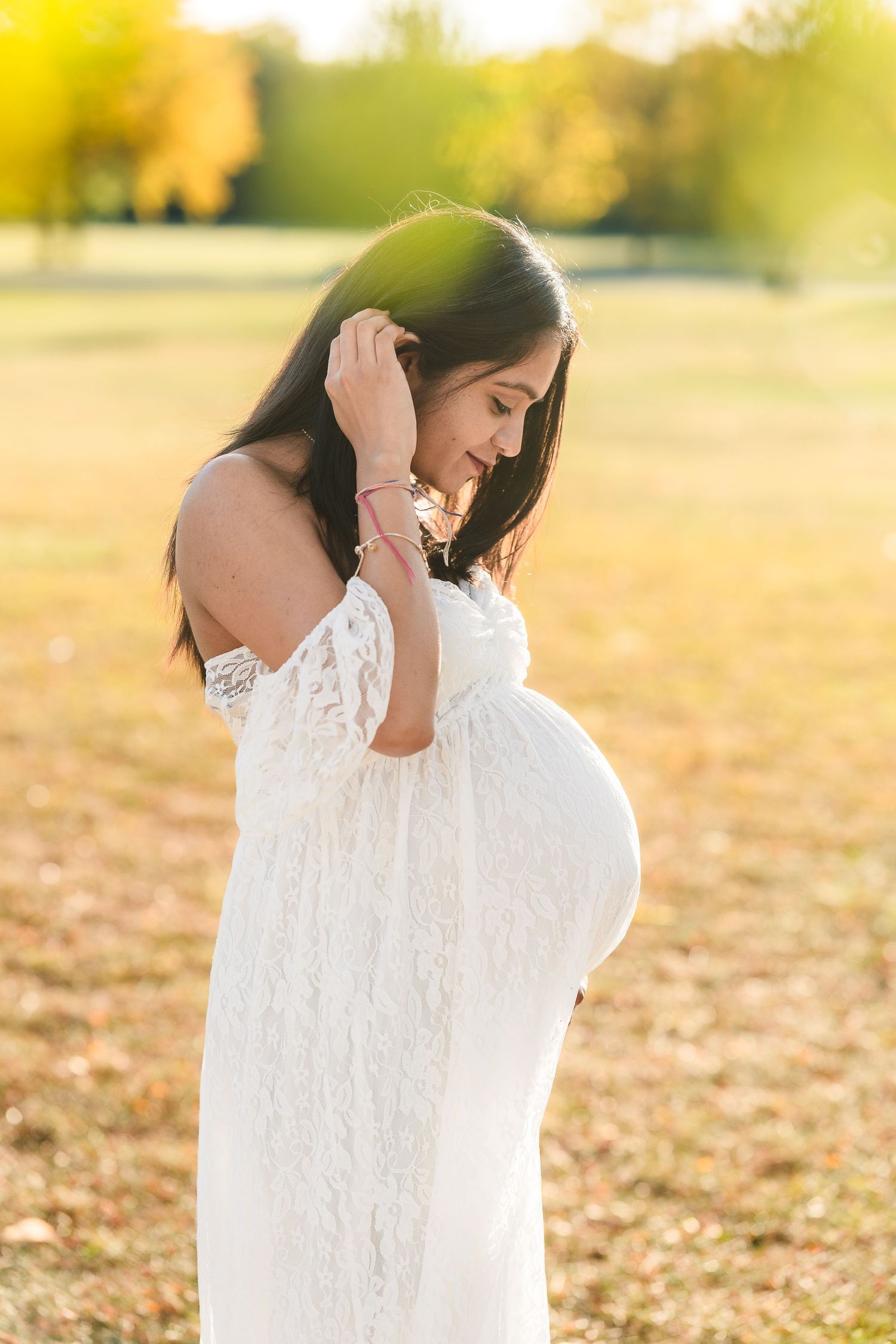 Side profile of expectant mother in white lace dress against golden sunset background.