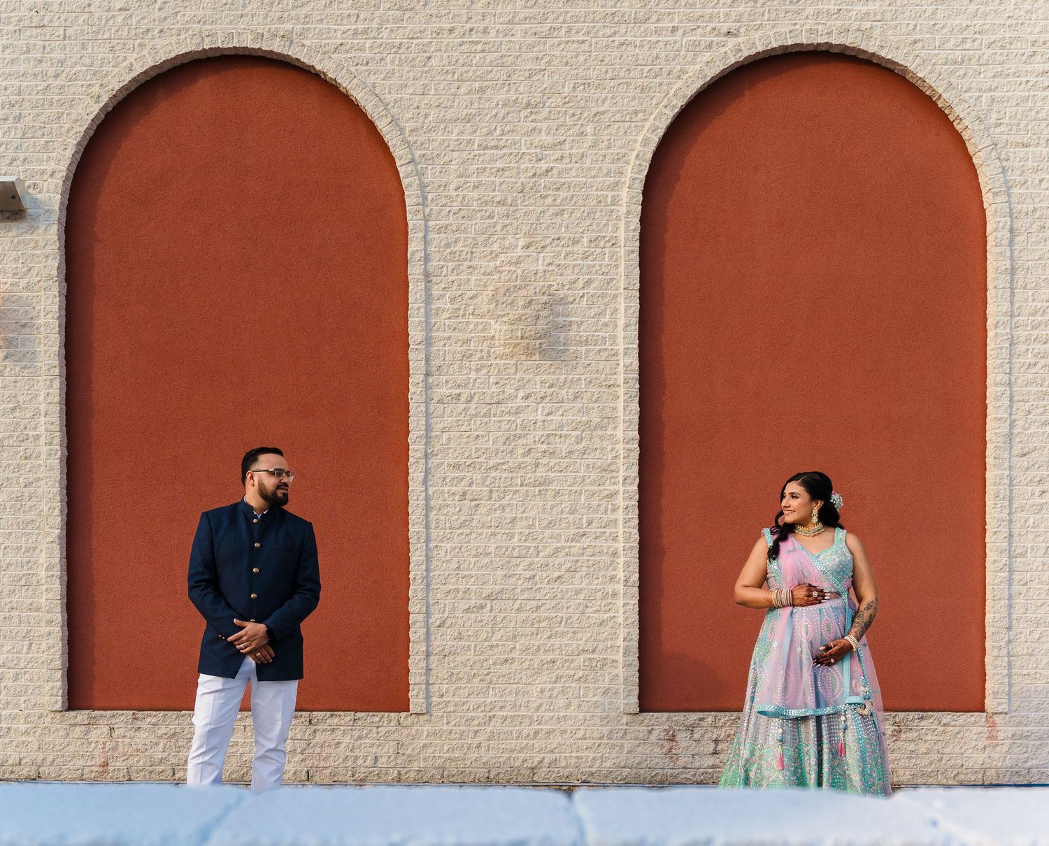 A couple stands separately in terracotta arched alcoves wearing coordinated formal attire.