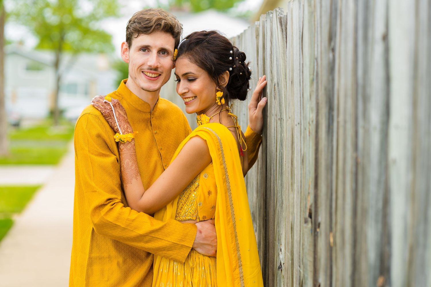 Couple in matching yellow outfits embrace near wooden fence outdoors.