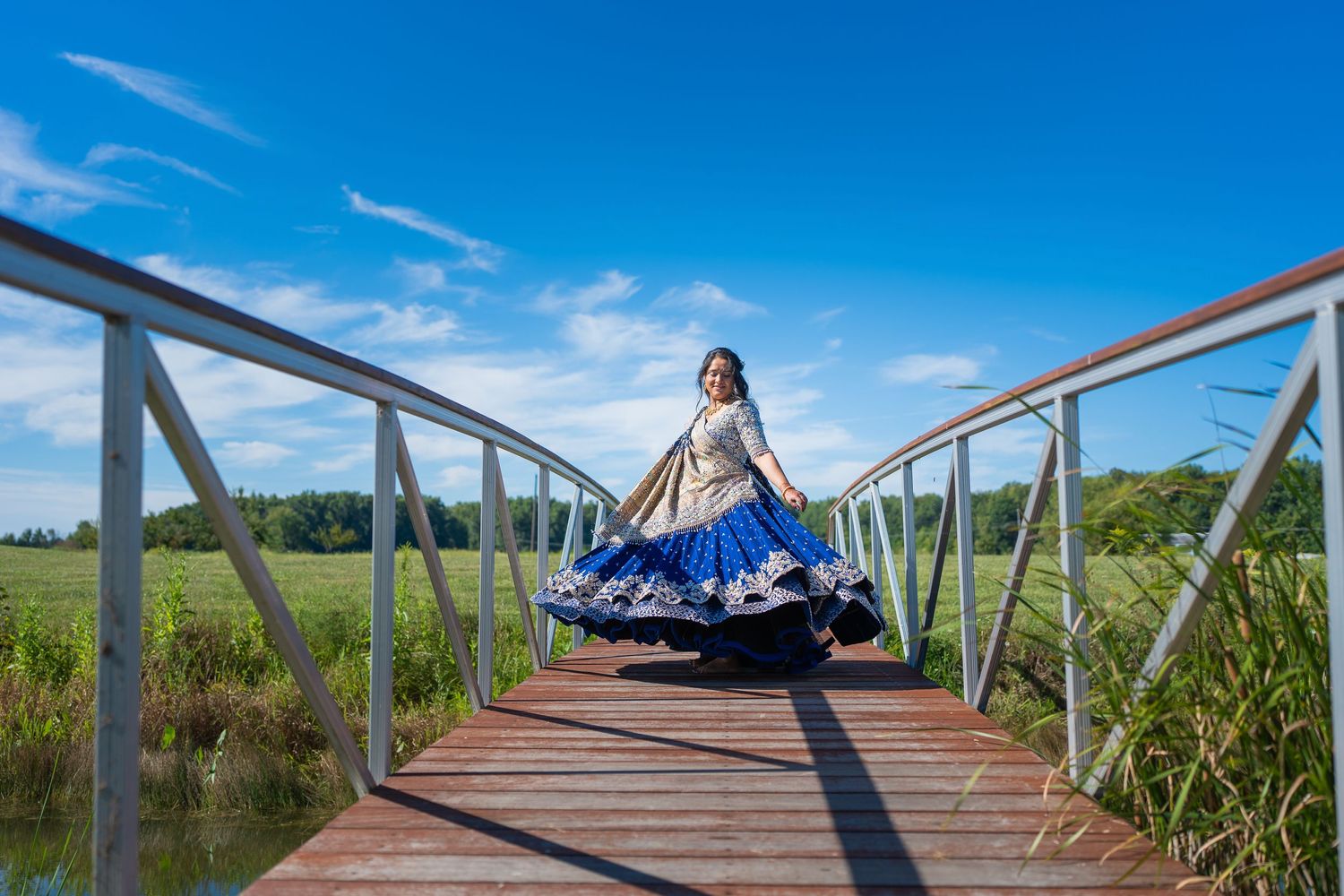 Person in blue traditional dress twirling on wooden bridge over marshland under bright blue sky.