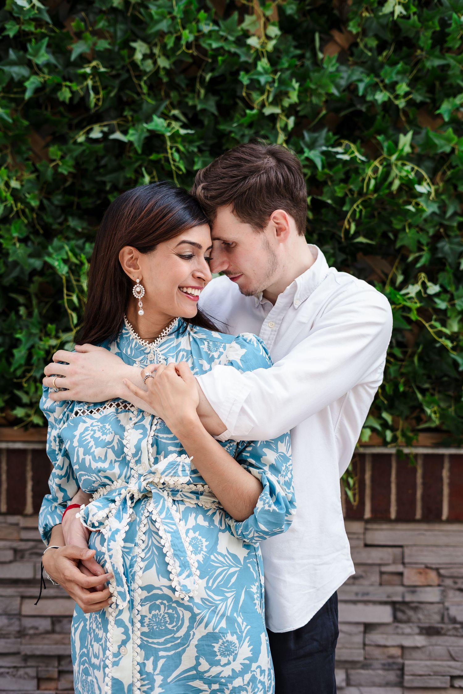 Romantic couple embraces against a green hedge backdrop, with her wearing a blue and white patterned dress.
