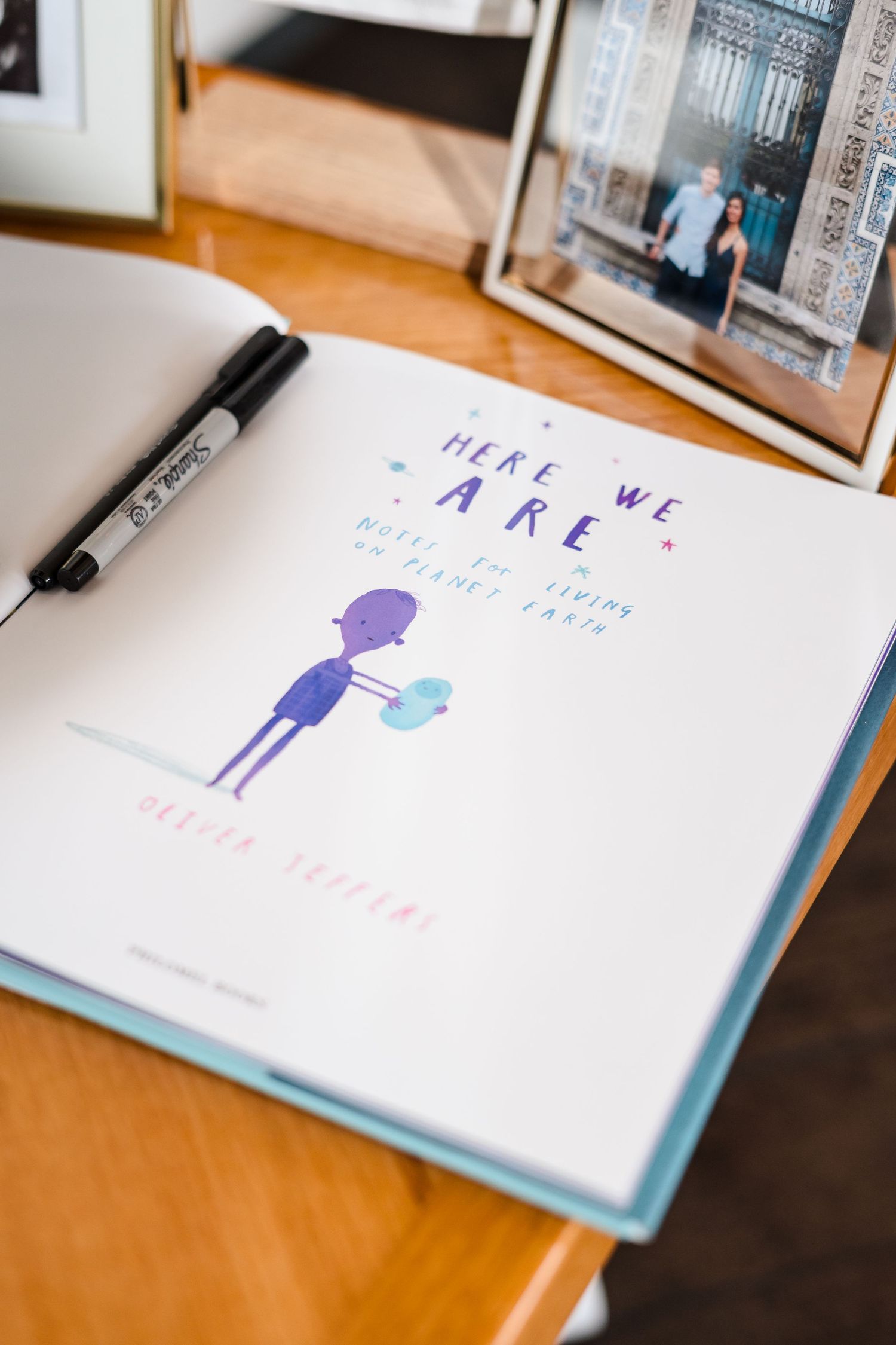 A book page with 'Here we are' text and a simple purple illustration of a figure holding something blue on a wooden desk.
