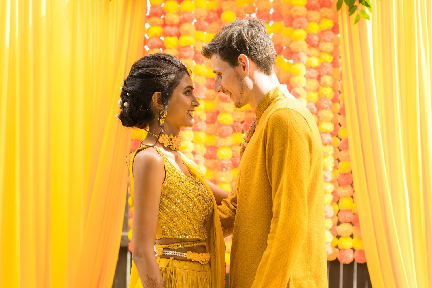 Couple in yellow and formal attire against vibrant orange and yellow draped backdrop with floral decorations.