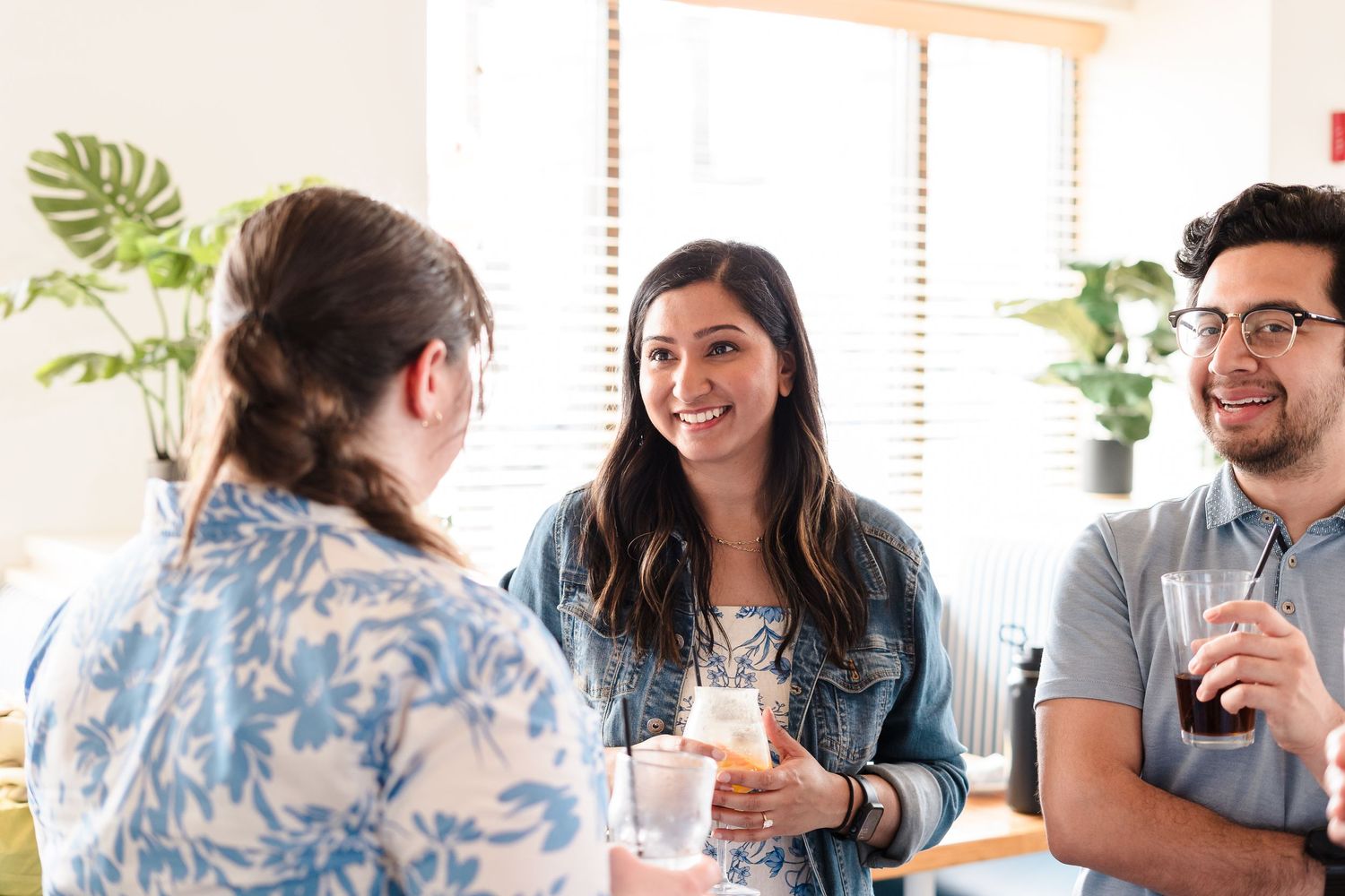 Three people enjoying drinks and conversation in a bright living room with houseplants and natural light.