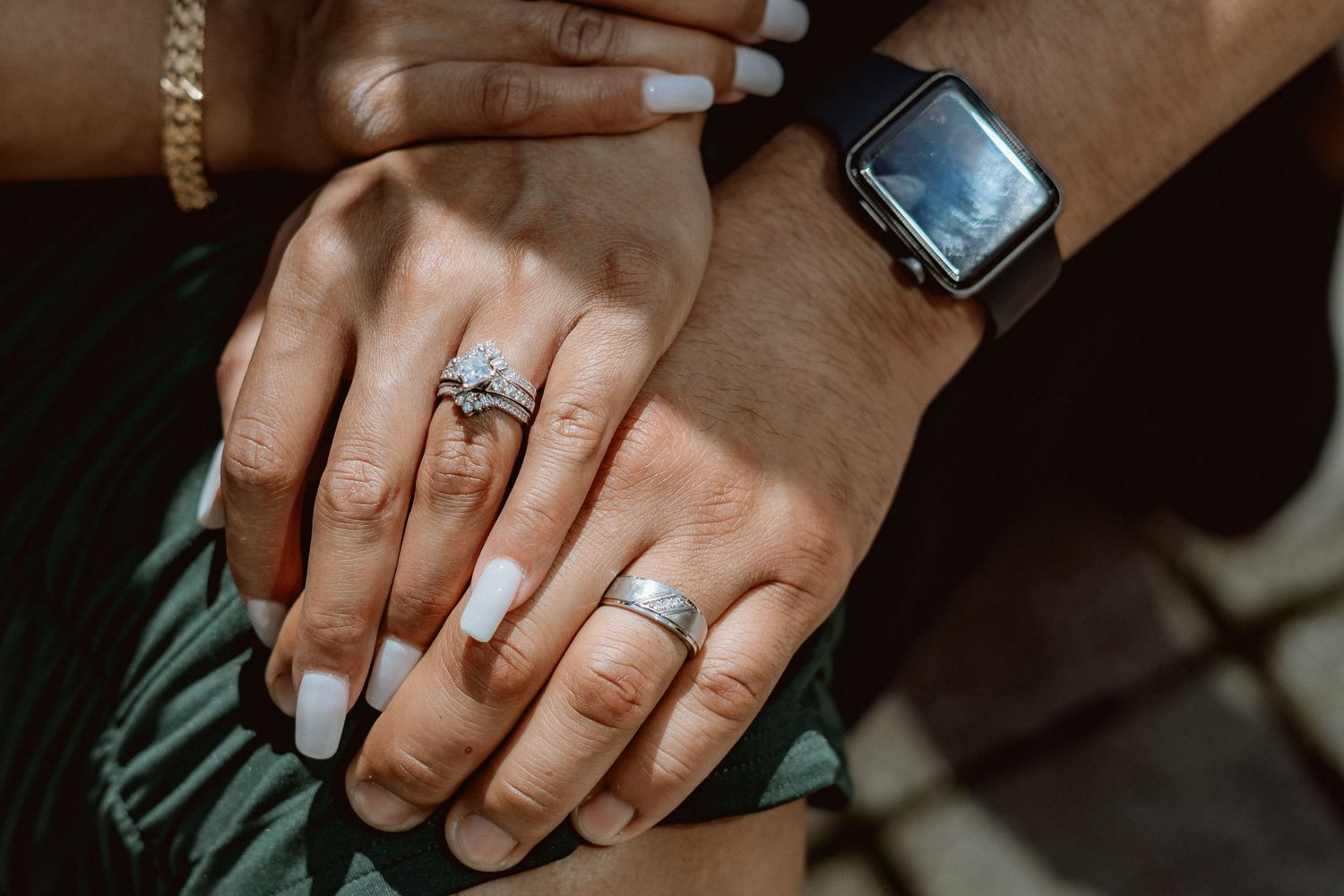 Close-up of hands with engagement rings and a smart watch against a dark background.