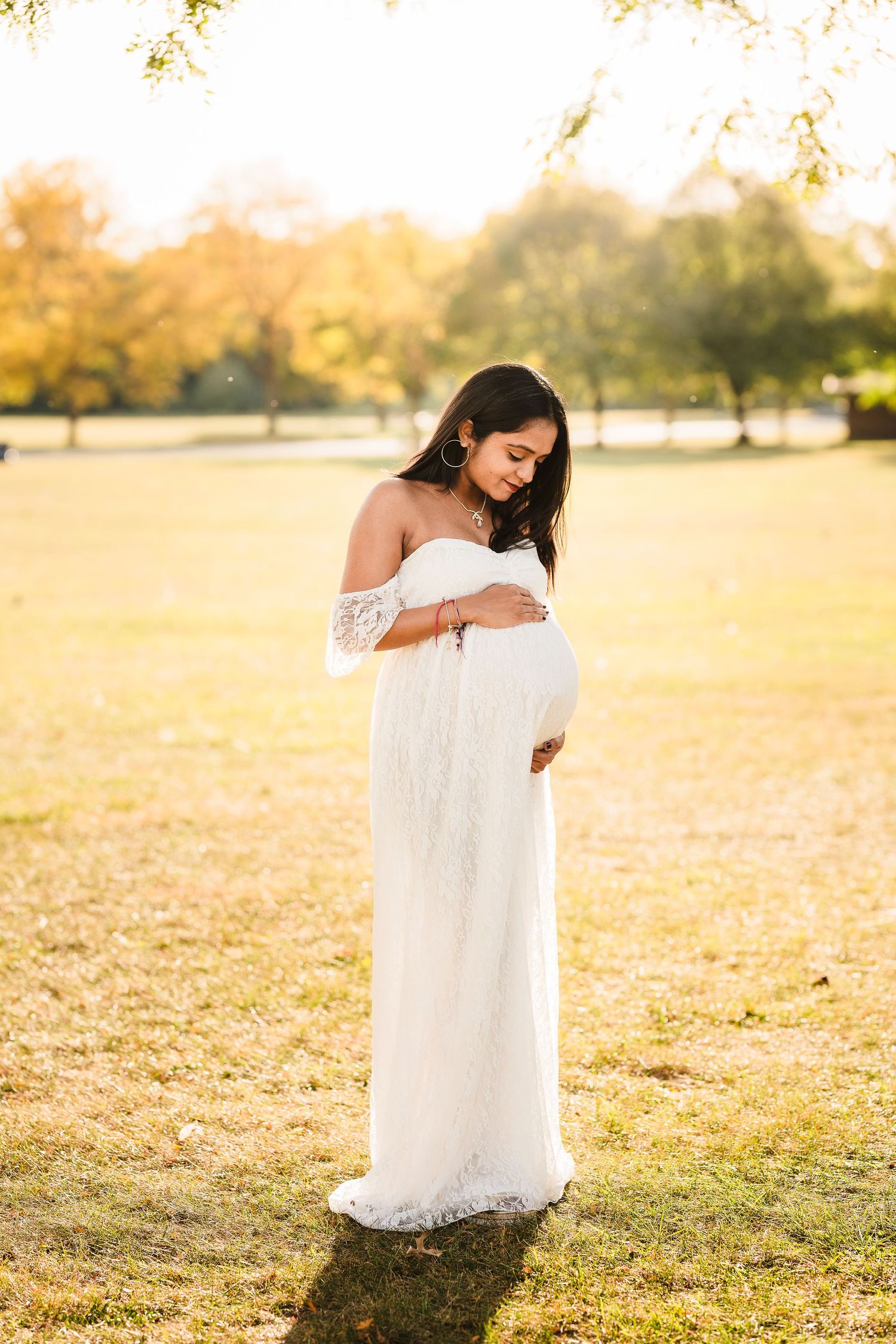 An expectant mother in a white off-shoulder dress poses in a sunlit field during golden hour.