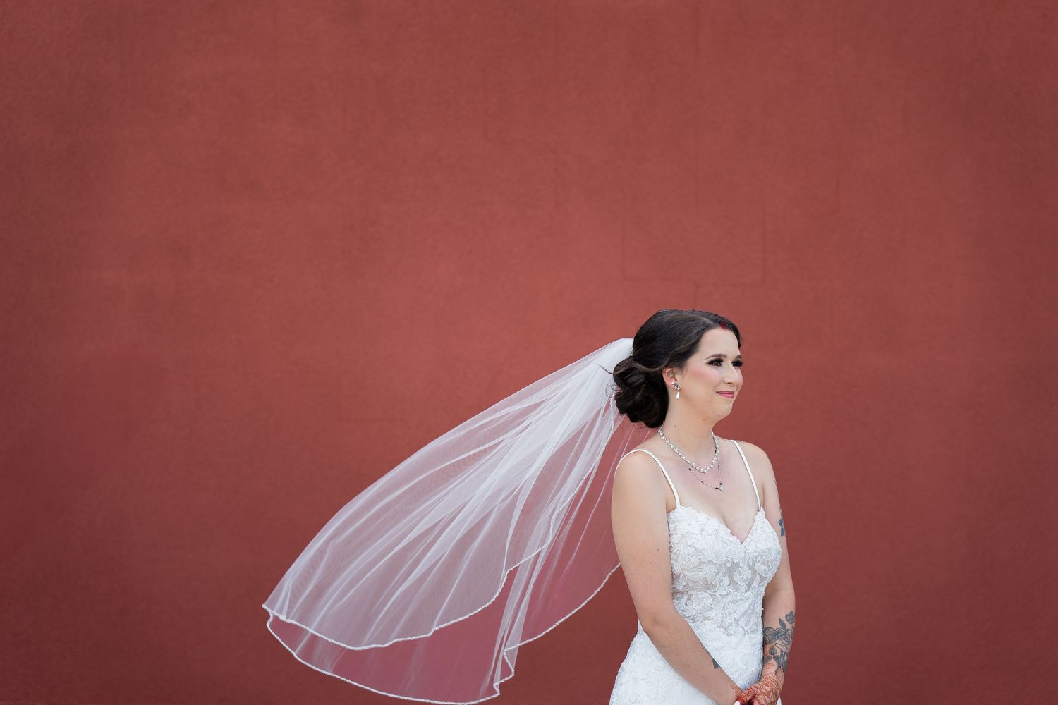 A bride in a white lace gown with flowing veil poses against a rustic red wall.
