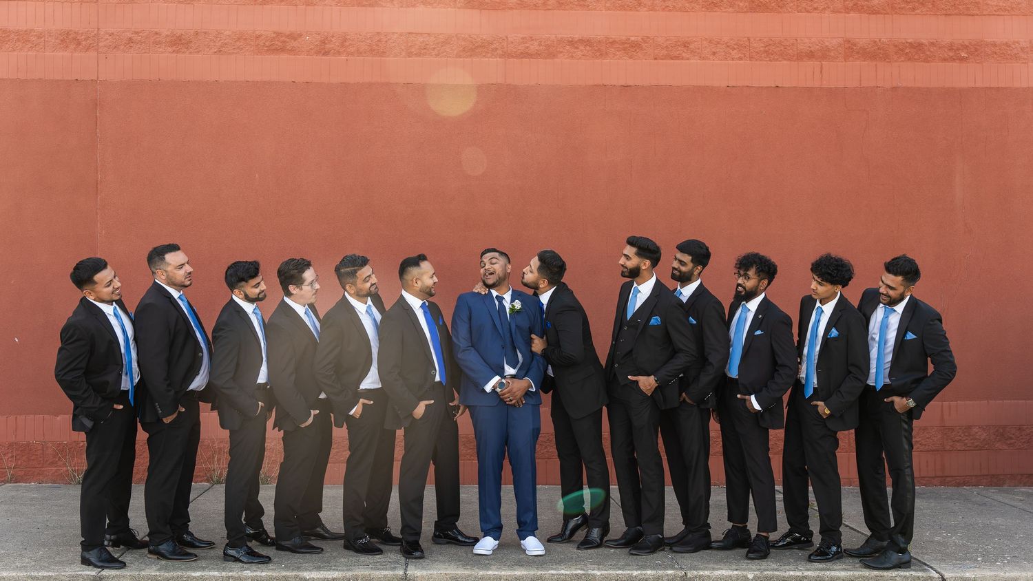 Groomsmen in dark suits line up against a terracotta colored wall in a stylish wedding party photo.