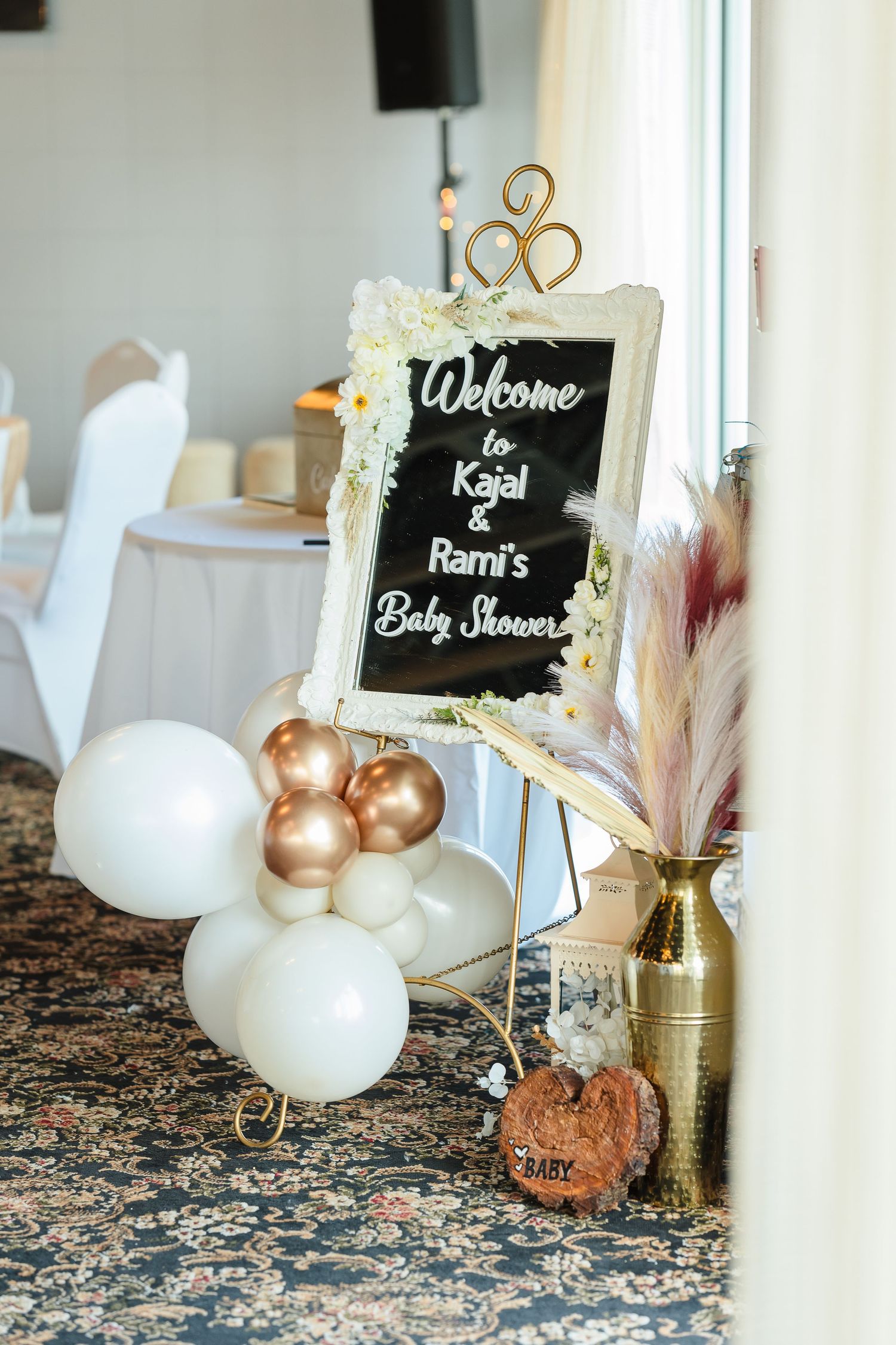 Decorative baby shower welcome sign with white and gold balloons and dried pampas grass accents.
