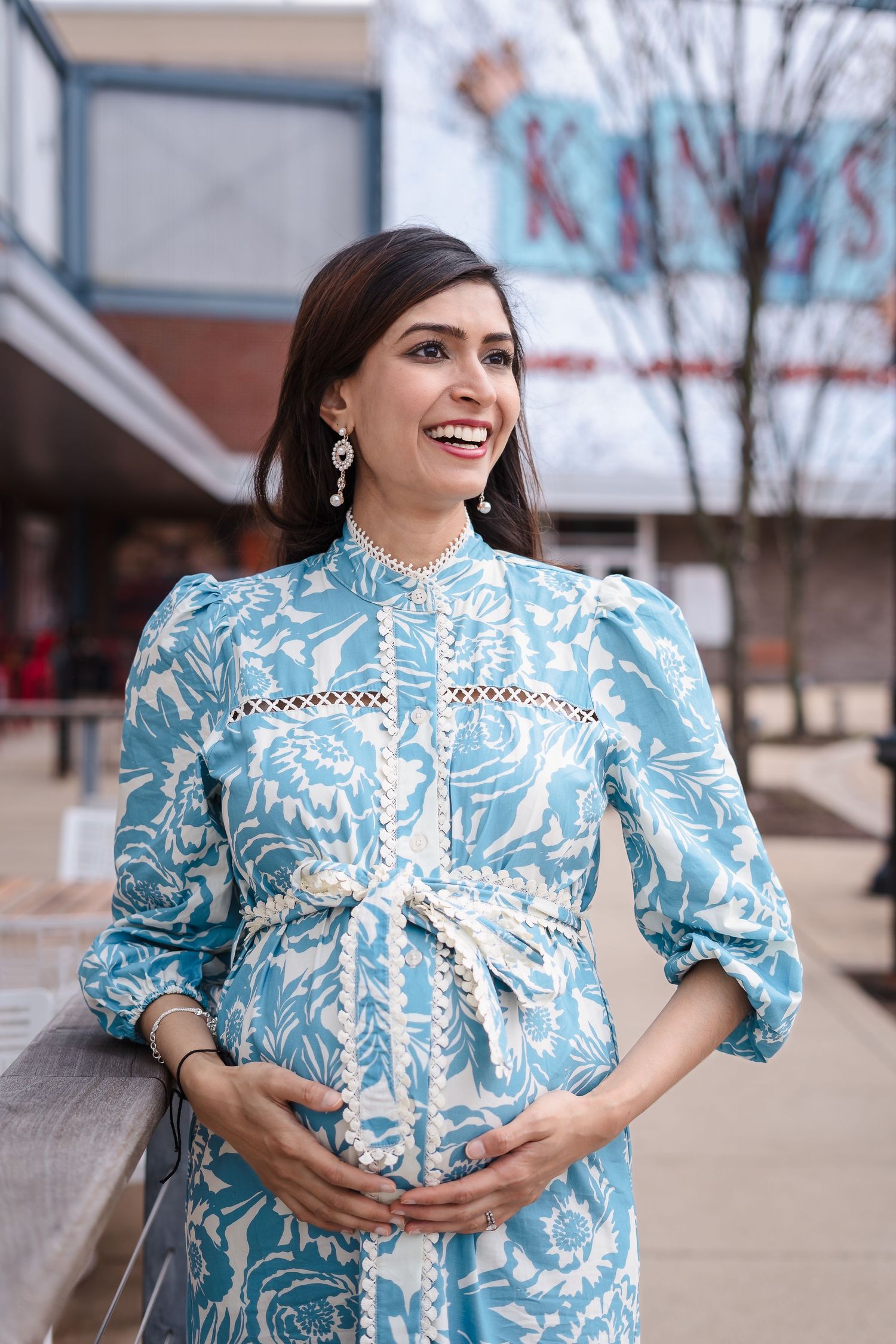 Pregnant woman wearing elegant light blue and white floral dress with balloon sleeves stands smiling outdoors.