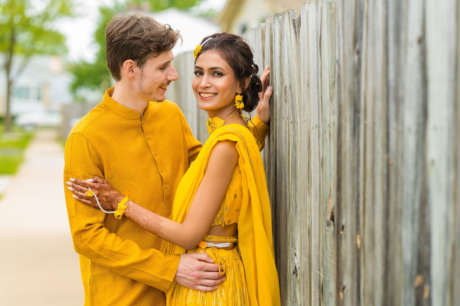 Couple in coordinating yellow attire share intimate moment against rustic fence.