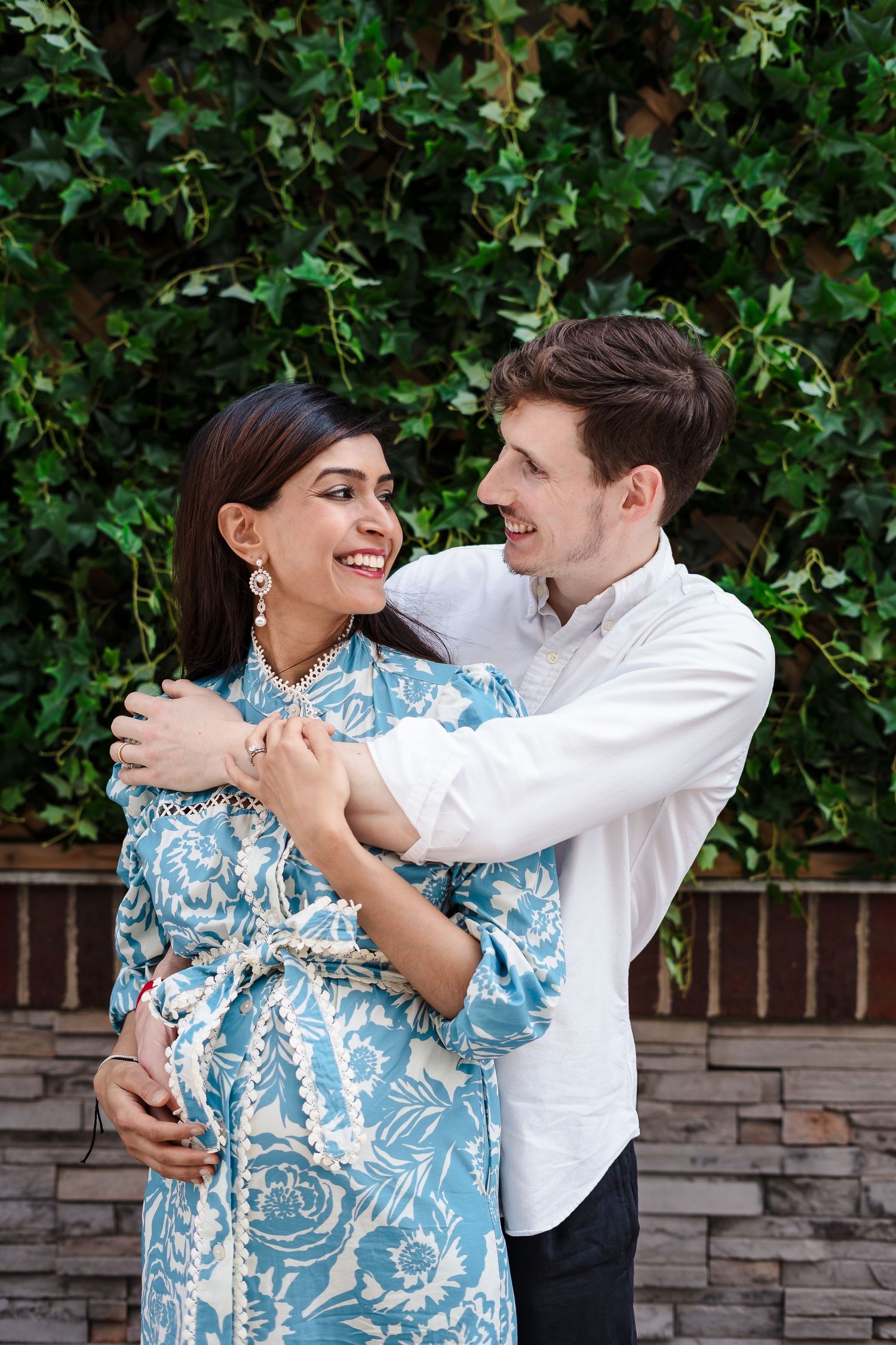 A couple shares a tender embrace in front of a lush green hedge, with the woman wearing a blue and white patterned dress.