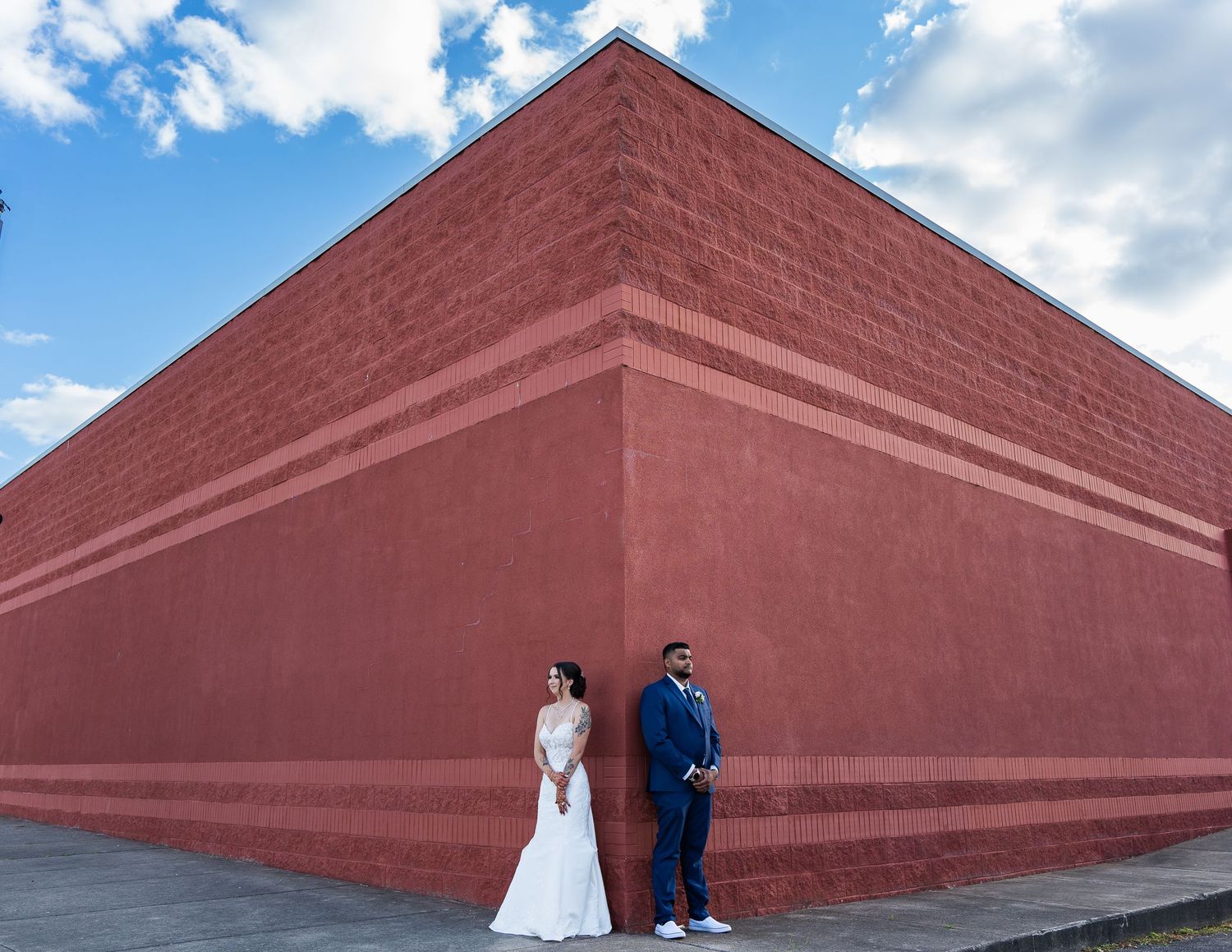 A couple in wedding attire poses against a striking red geometric wall under blue skies.