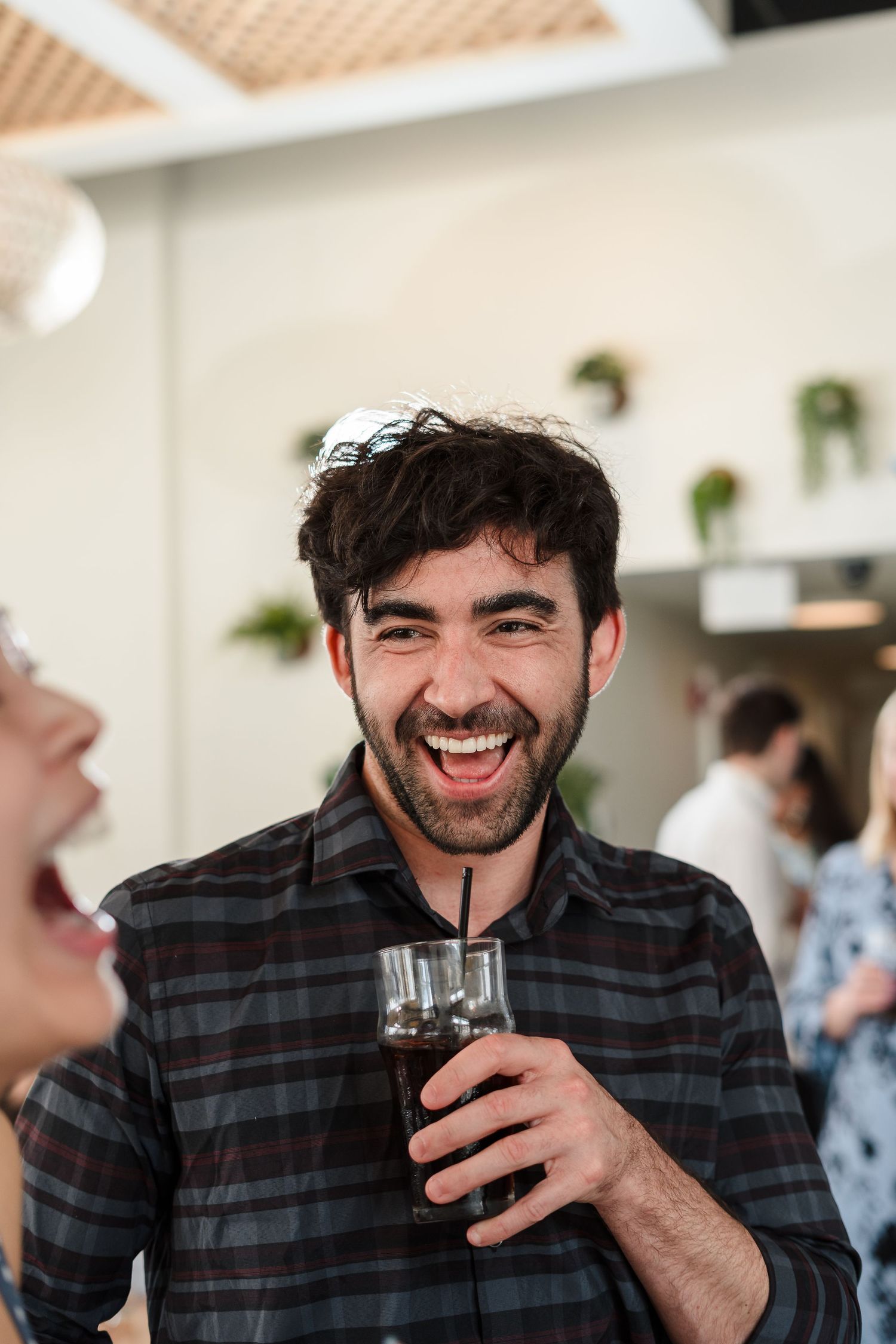 Someone laughing while holding a drink at a social gathering with houseplants visible in the background.