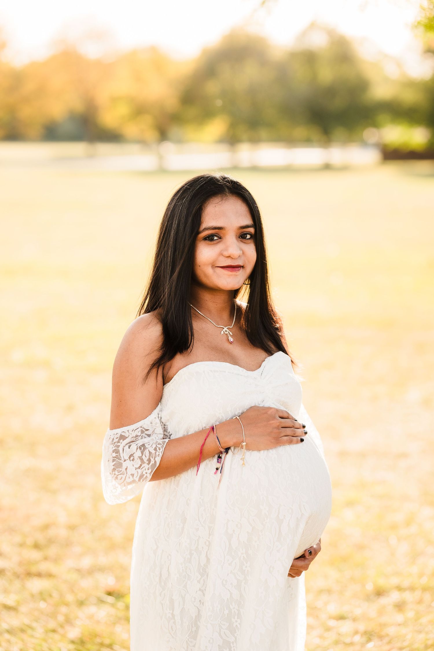 Woman in white lace dress poses in golden autumn field during maternity photoshoot.
