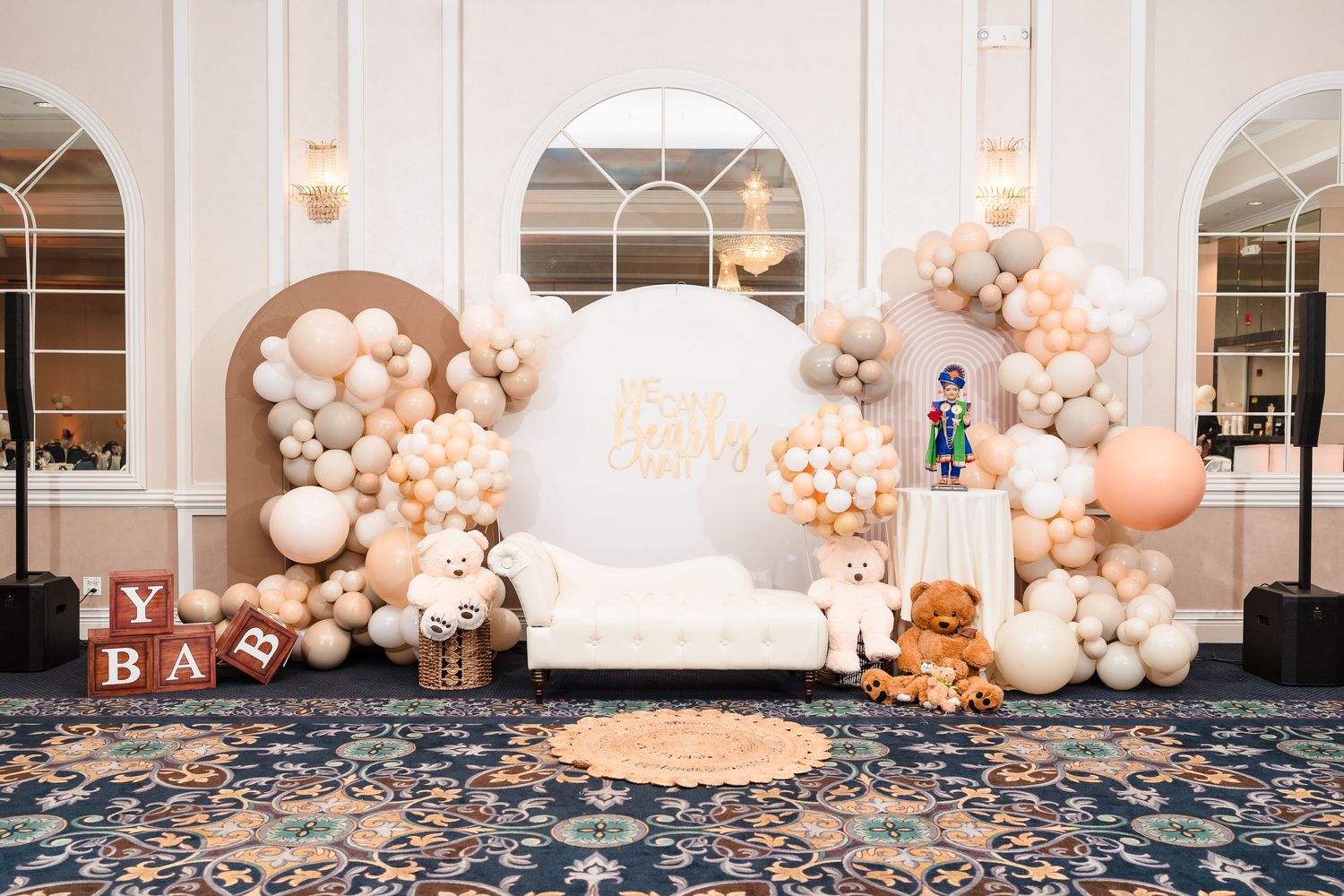 Elegant baby shower setup with white sofa and blush balloon arch against white arched walls.