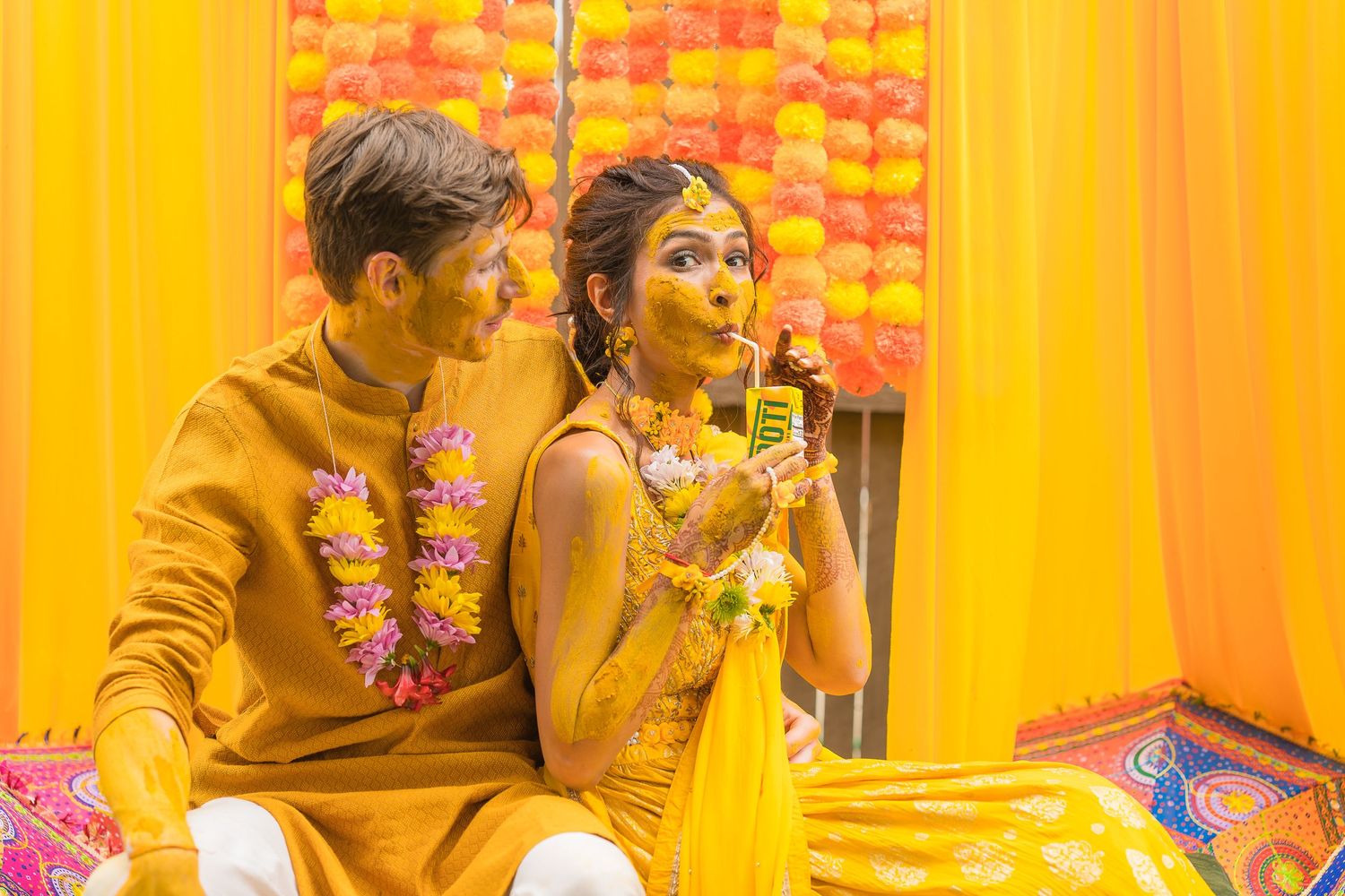 Couple in traditional yellow Indian attire sitting together amid colorful decorations.