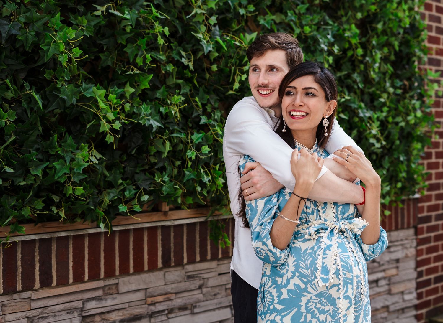 A joyful couple poses together against a brick wall and green hedge backdrop.