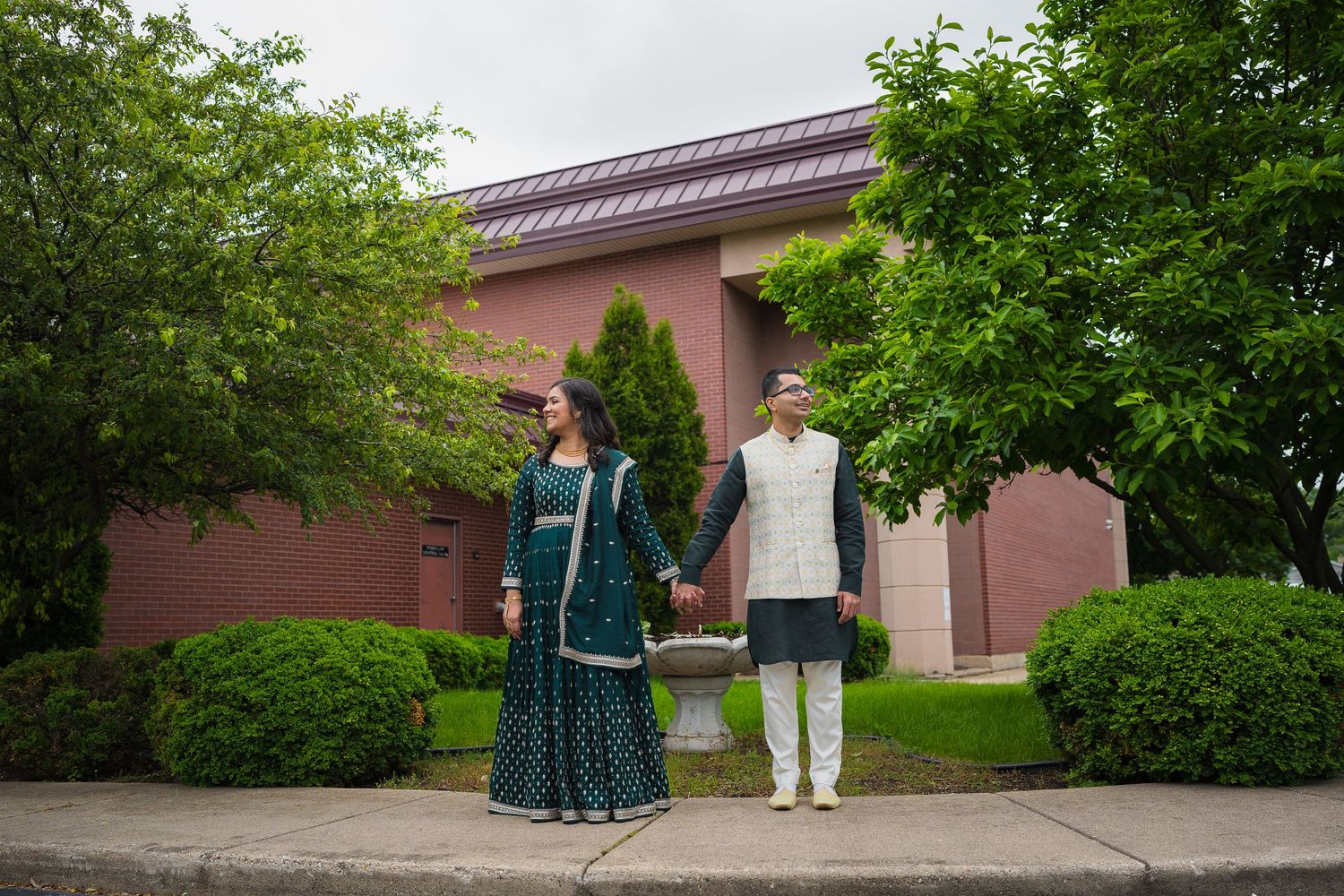 A couple in traditional Indian attire poses in front of a pink building surrounded by greenery.