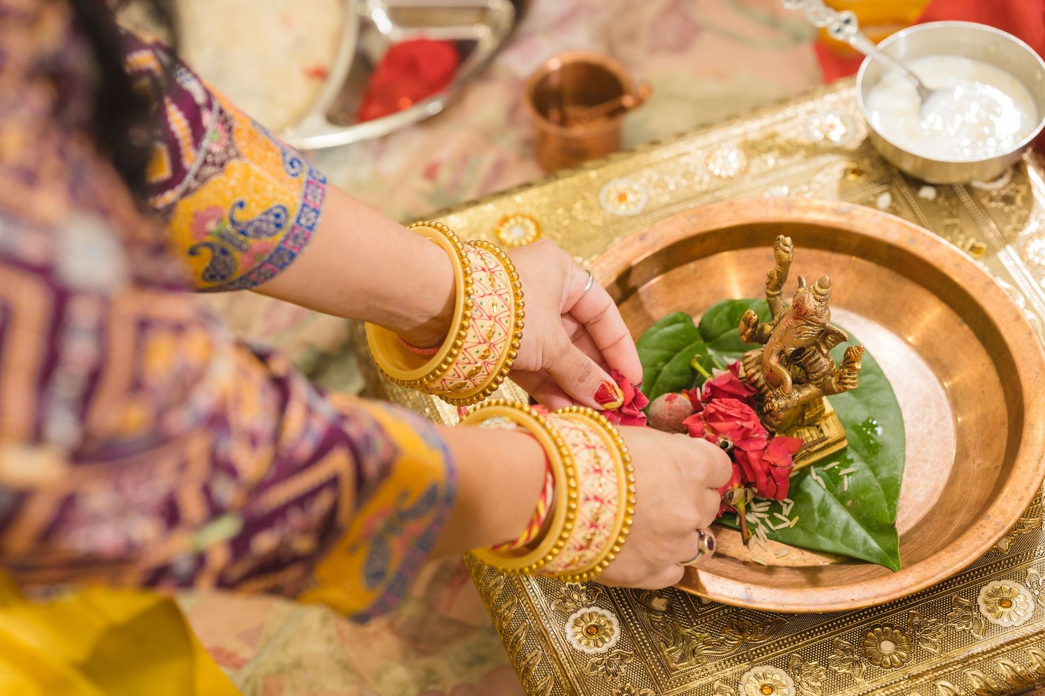Hands adorned with gold bangles reaching for traditional Indian food items on a decorative plate.