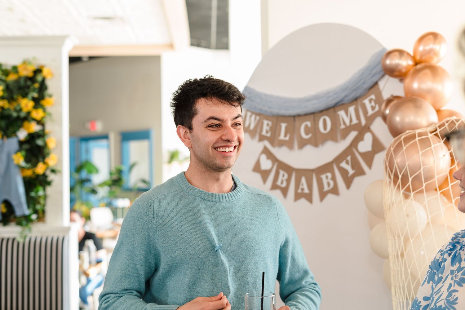 Festive party scene with teal sweater, welcome banner and gold balloon decorations in bright indoor setting.