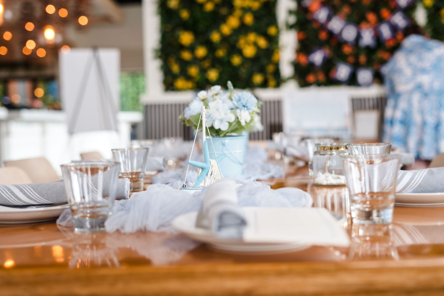 Elegant wedding table setting with glassware, plates and blue floral centerpiece against blurred festive backdrop.