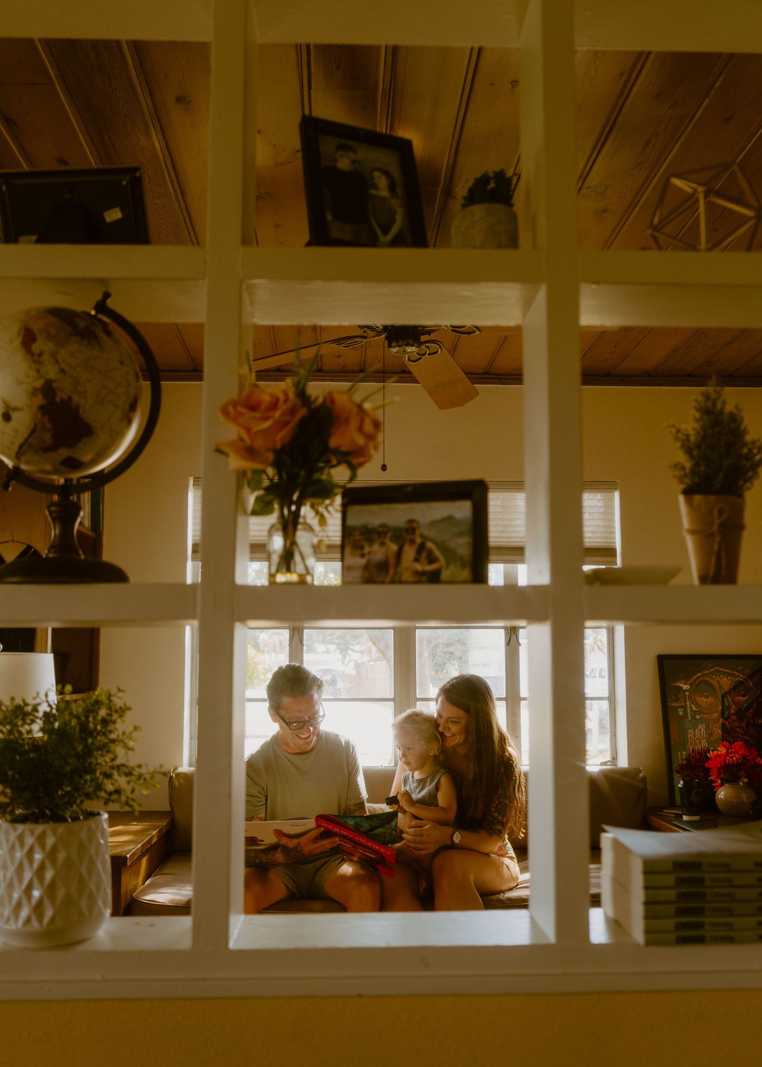 Family reading together in a cozy, warmly lit room with shelves, globe, and flowers visible through a white grid window frame.