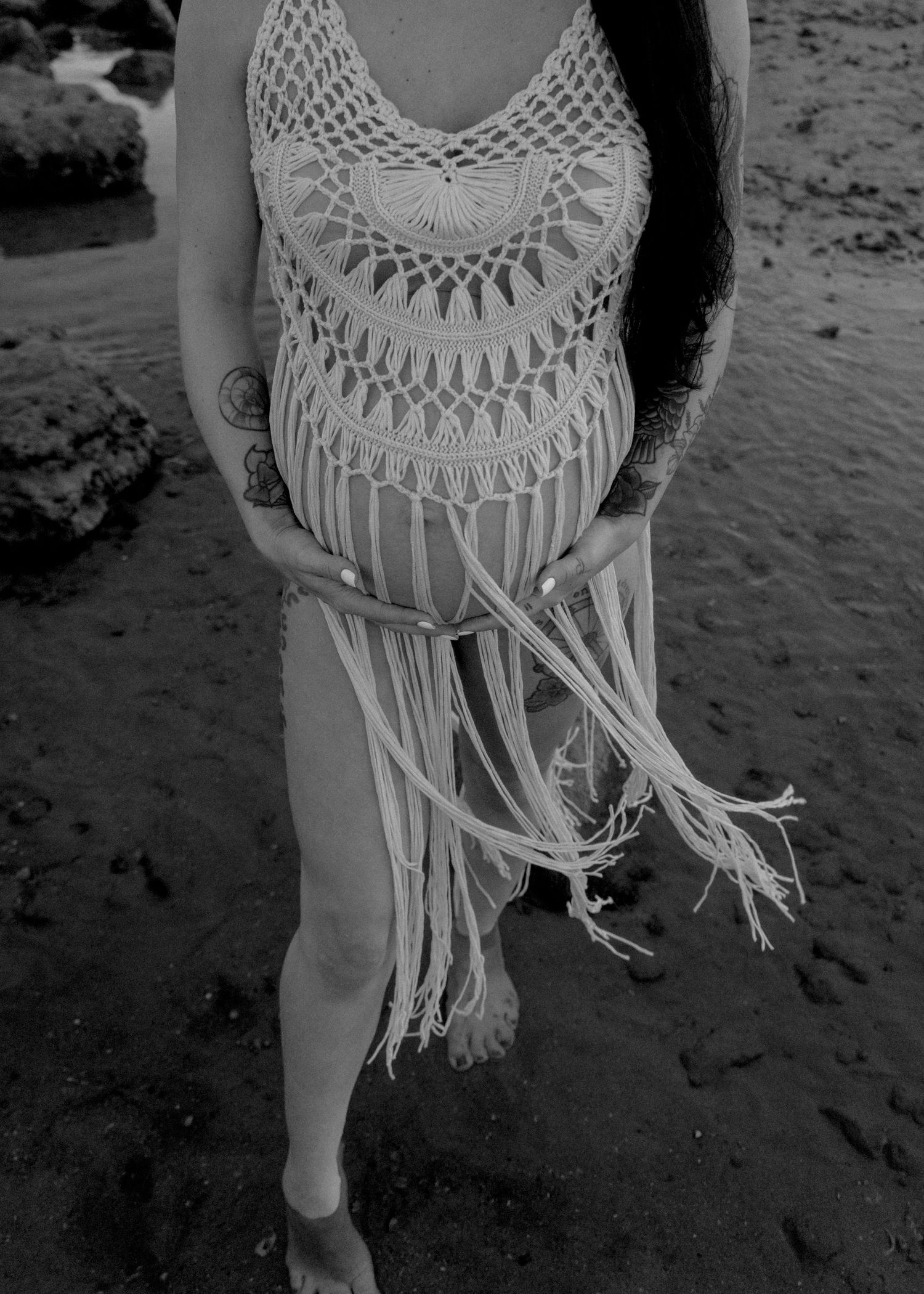 Pregnant woman in crochet fringe dress cradling baby bump on rocky beach in black and white photo.