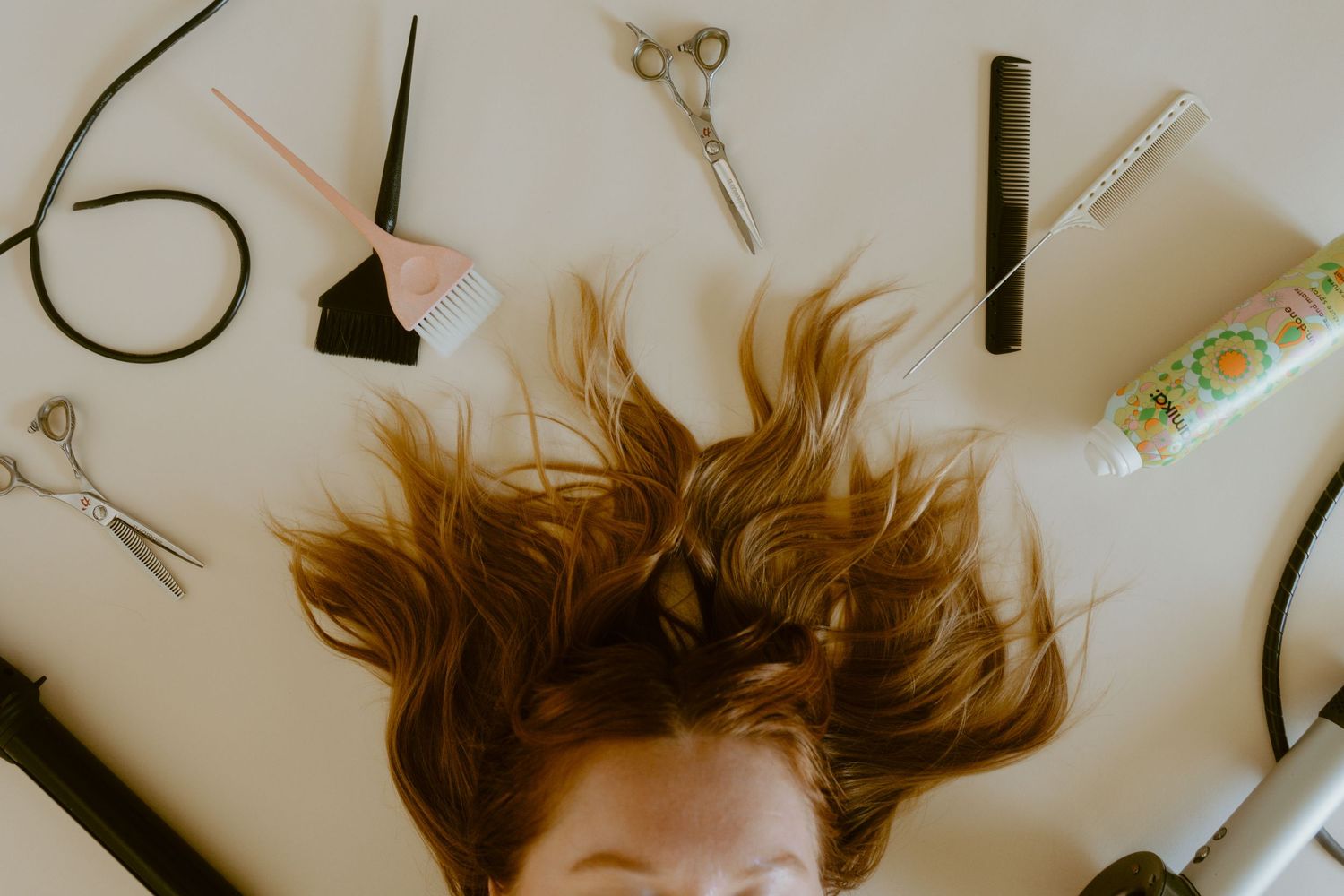 Woman lying on white surface surrounded by hair styling tools including scissors, combs, and spray bottle.